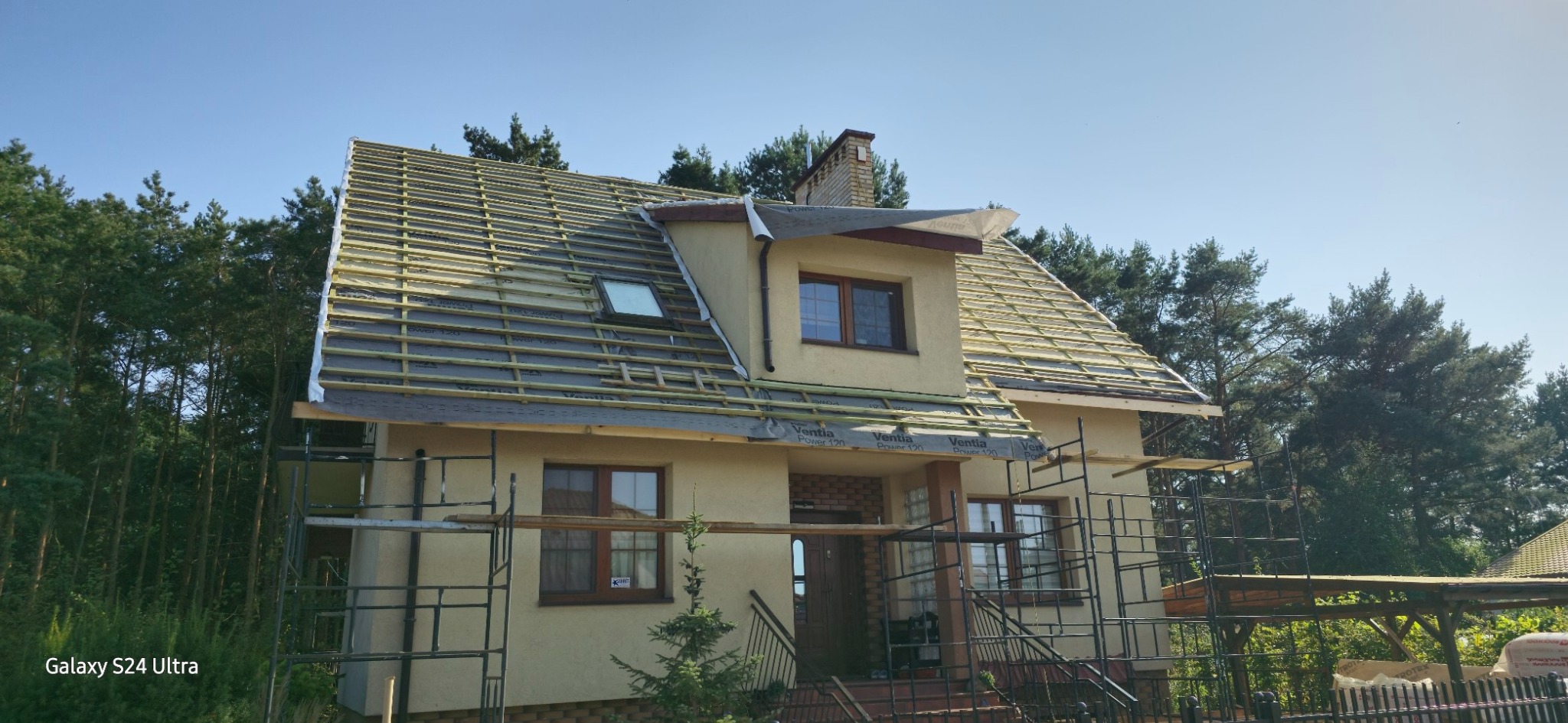 Dom jednorodzinny w trakcie budowy dachu, widoczne łaty, membrana dachowa i rusztowania, tło stanowią drzewa.