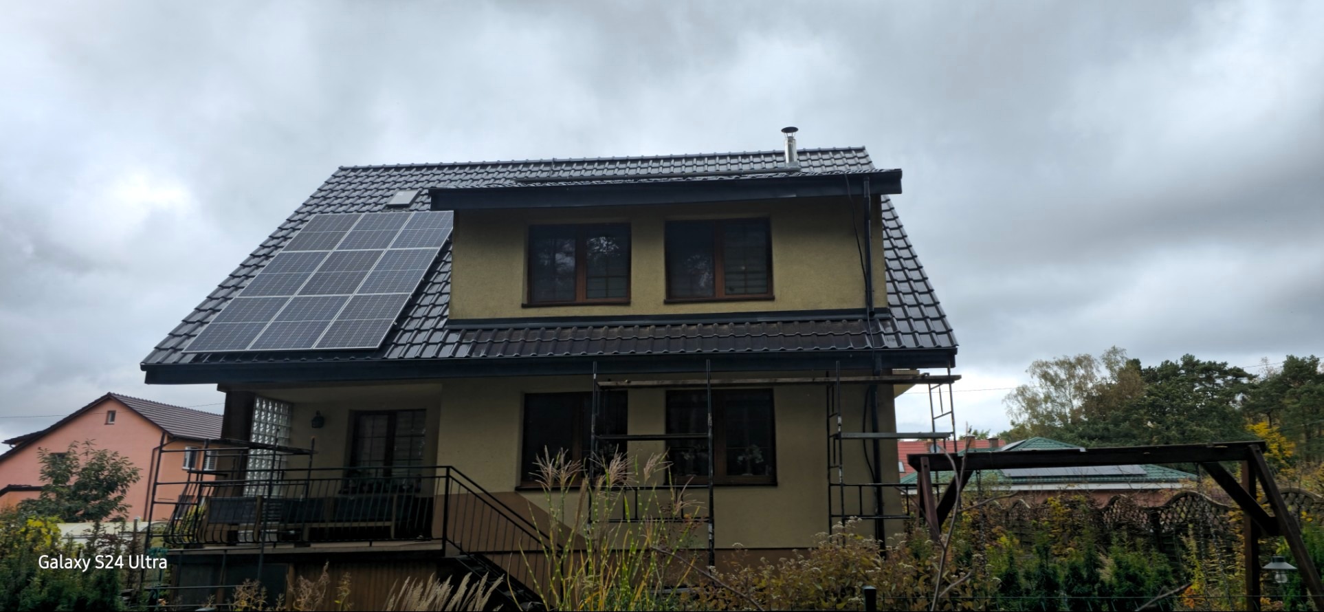 Dom jednorodzinny z ciemną dachówką i panelami słonecznymi na dachu, widoczne rusztowanie przy elewacji, pochmurne niebo w tle.