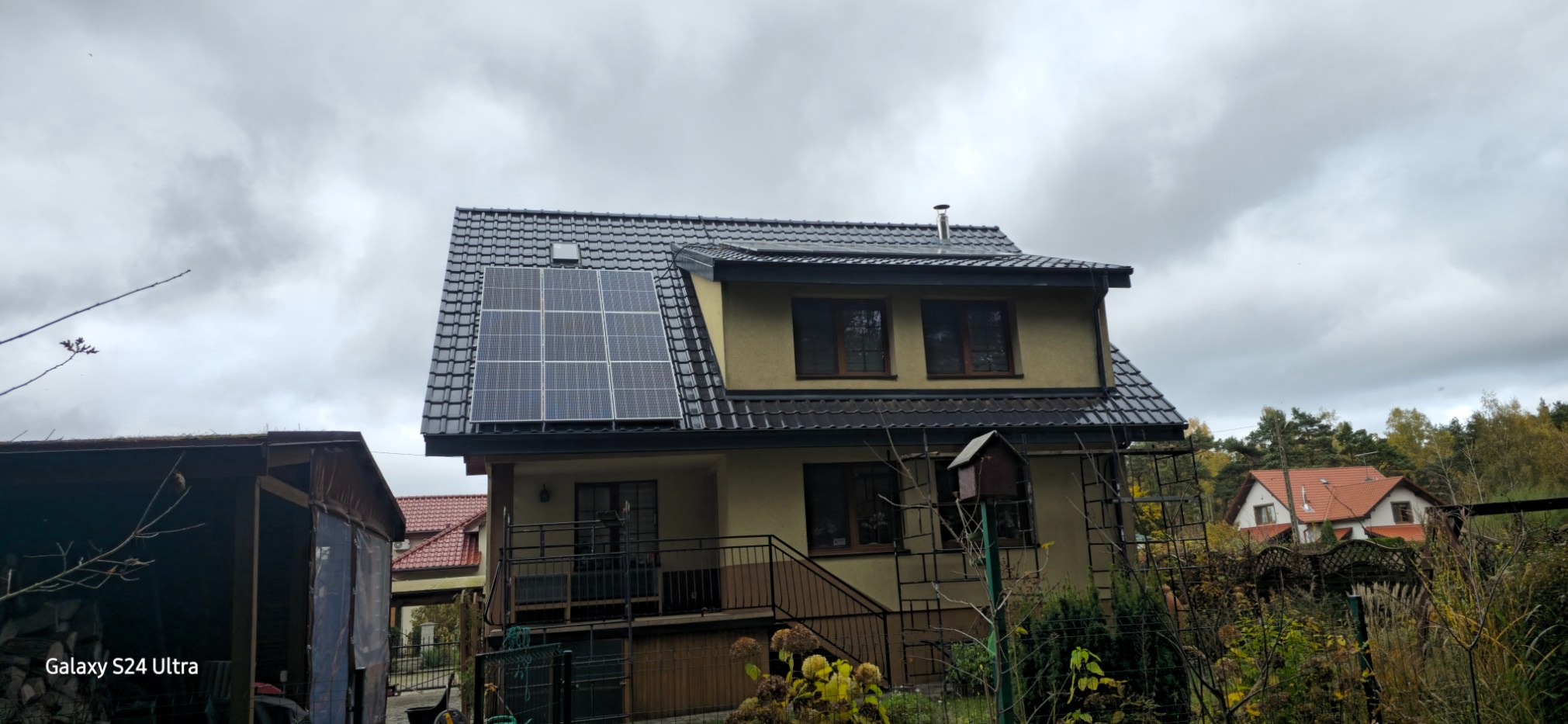 Dom jednorodzinny z nowym, ciemnym dachem pokrytym dachówką i panelami słonecznymi, widok z ogrodu w pochmurny dzień.