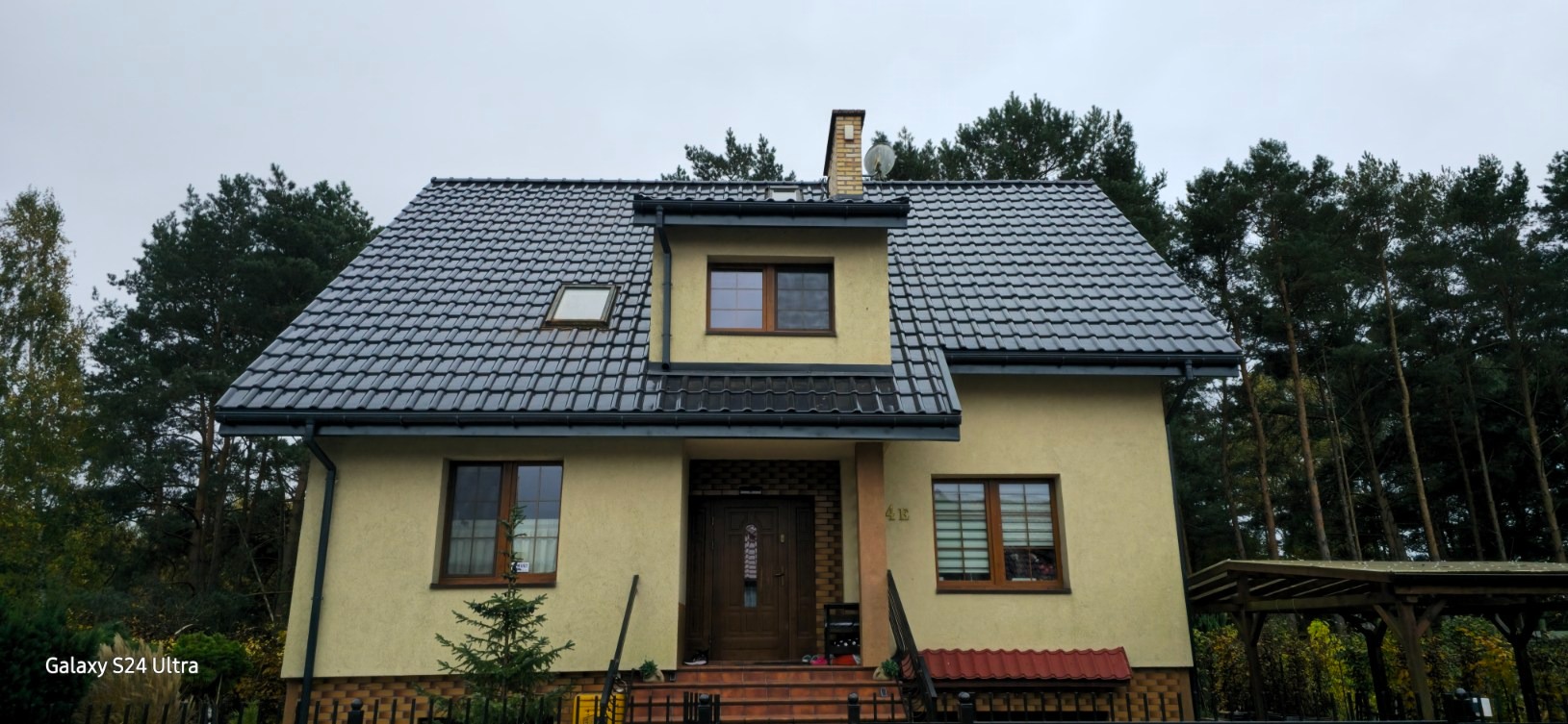 Dom jednorodzinny z nowym, ciemnoszarym dachem z dachówki ceramicznej, widoczny komin i okno dachowe, elewacja w kolorze jasnego beżu, otoczony drzewami iglastymi.