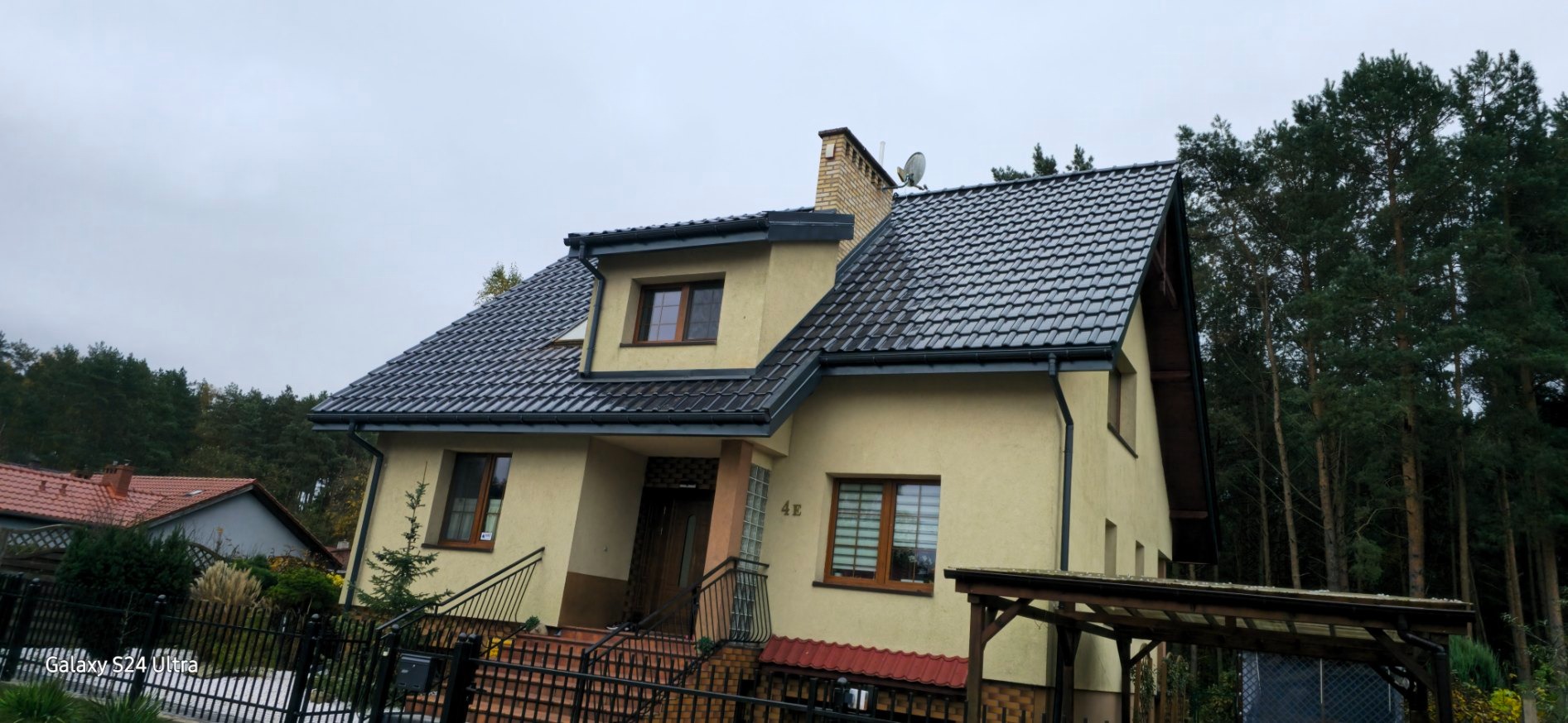 Dom jednorodzinny z nowym, błyszczącym dachem z dachówki ceramicznej w kolorze grafitowym i odnowioną elewacją w odcieniach beżu, widoczny zza czarnego, metalowego ogrodzenia.