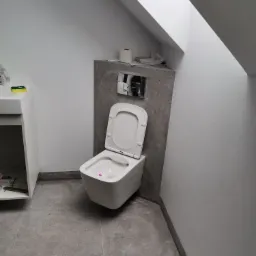 Nowoczesna toaleta podwieszana z otwartą klapą w łazience na poddaszu, widoczne szare płytki podłogowe i fragment szafki umywalkowej.