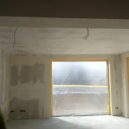 Pokój w trakcie prac wykończeniowych: ściany z nałożonymi warstwami gładzi, okna zabezpieczone folią malarską, widoczne wyprowadzone kable elektryczne.