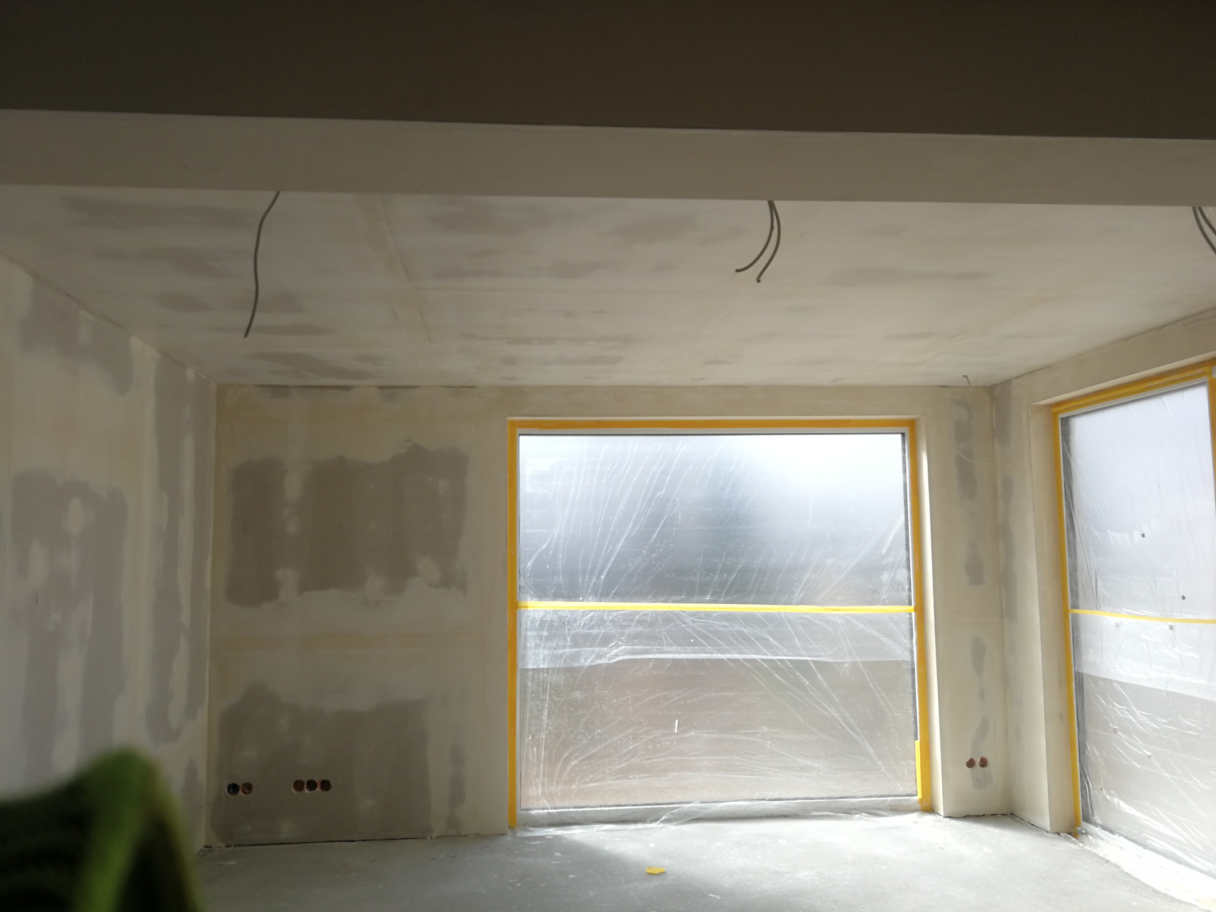 Pokój w trakcie prac wykończeniowych: ściany z nałożonymi warstwami gładzi, okna zabezpieczone folią malarską, widoczne wyprowadzone kable elektryczne.