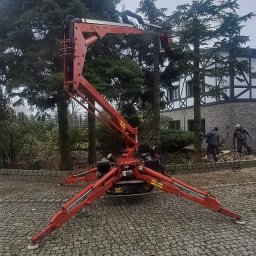 FIRMA USŁUGOWO-HANDLOWA DOMAC DOMINIKA PILARSKA MACIEJ GRZESIAK S.C. - Czerwony podnośnik koszowy na gąsienicach, rozstawiony na brukowanej powierzchni, z dwoma pracownikami w tle przycinającymi drzewa, widoczne ścięte gałęzie na ziemi.