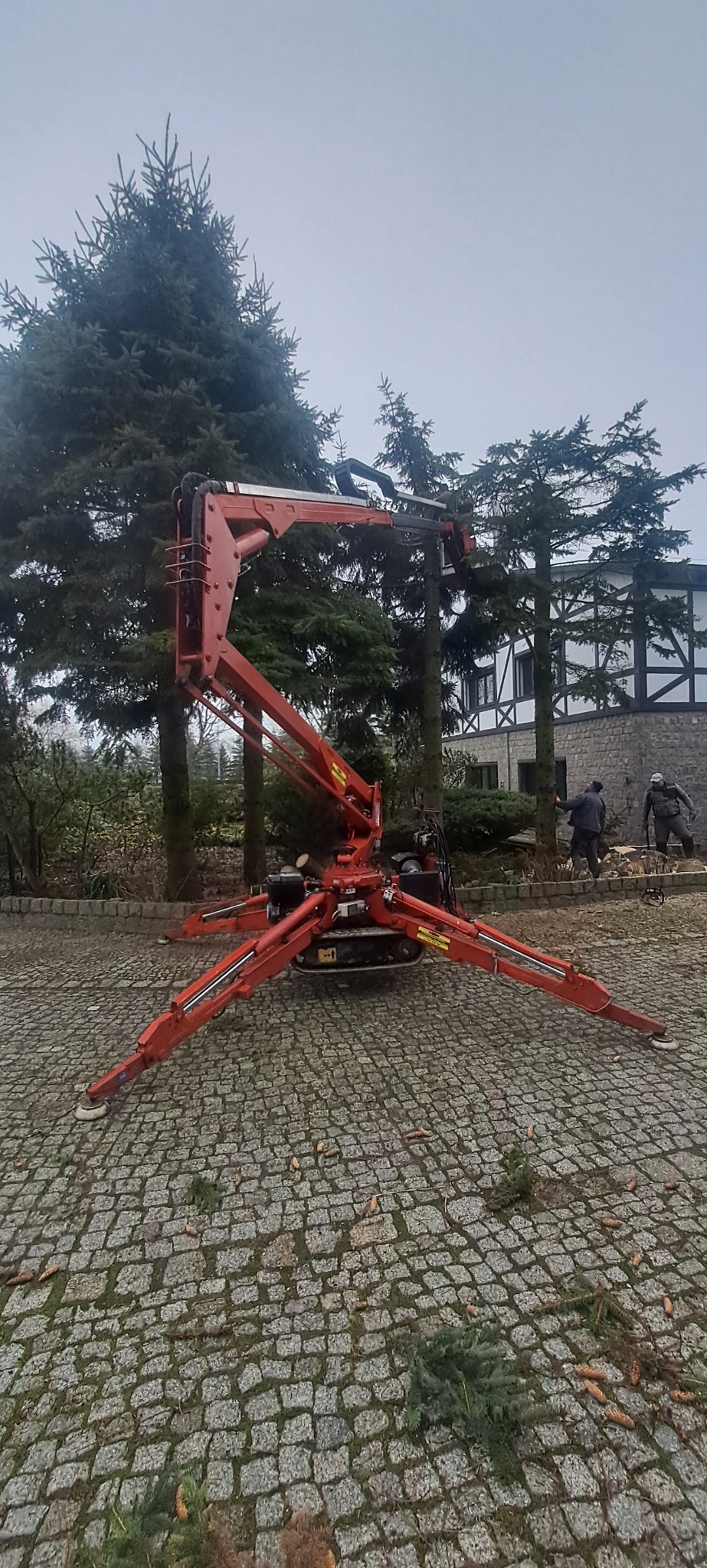 Czerwony podnośnik koszowy na gąsienicach, rozstawiony na brukowanej powierzchni, z dwoma pracownikami w tle przycinającymi drzewa, widoczne ścięte gałęzie na ziemi.