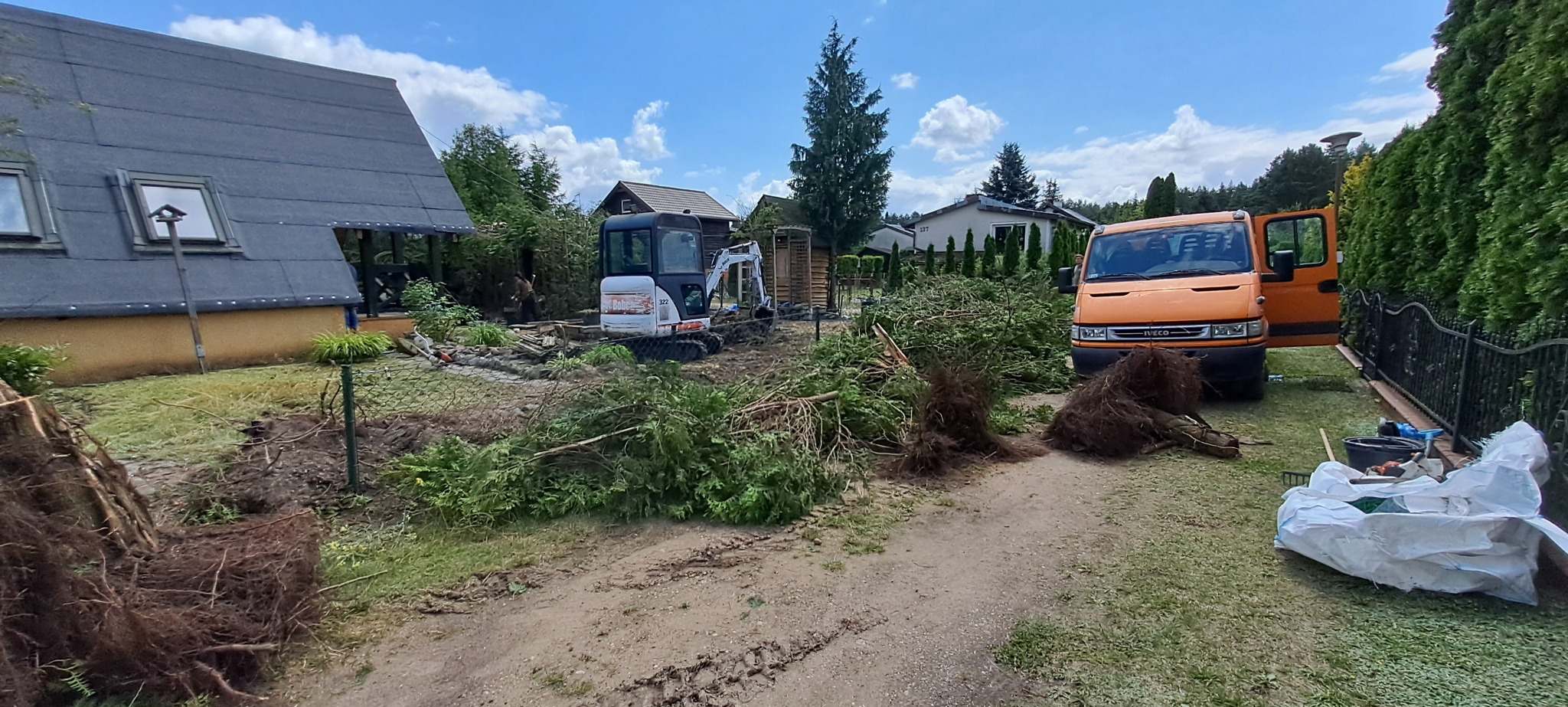 Teren po wycince drzew z widocznym mini-koparką, stertą gałęzi i pomarańczowym samochodem dostawczym na tle domów.