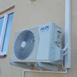 Zewnętrzna jednostka klimatyzacji AUX DC Inverter zamontowana na elewacji budynku z widocznymi rurami i osłoną instalacji.