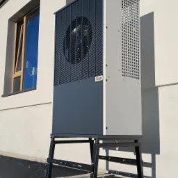 Zewnętrzna pompa ciepła powietrze-woda, model stojący na betonowych fundamentach przy białej ścianie budynku z widocznym oknem i niebieskim niebem w tle.