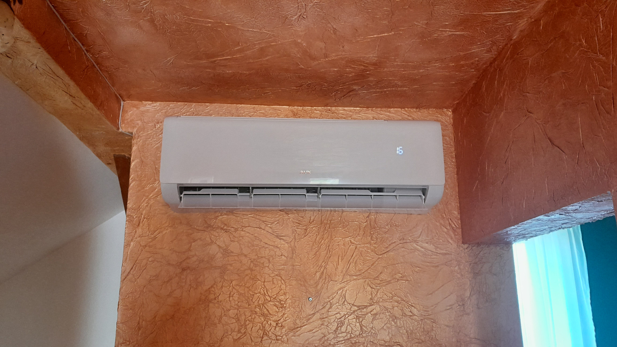 Biały klimatyzator marki AUX zamontowany na ścianie o fakturze imitującej skórę, z widocznym wyświetlaczem temperatury ustawionym na 16 stopni Celsjusza.