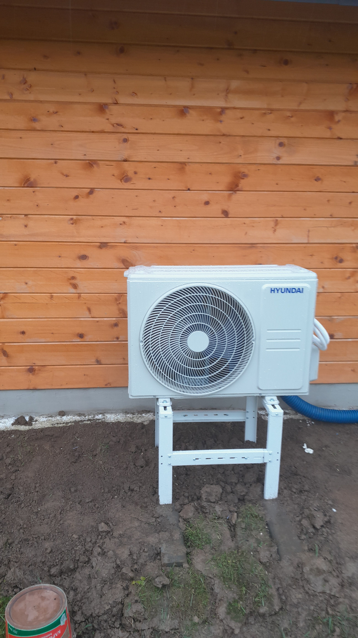 Biała jednostka zewnętrzna klimatyzacji Hyundai, zamontowana na metalowym stelażu, z niebieskim przewodem, na tle drewnianej ściany budynku w trakcie instalacji.