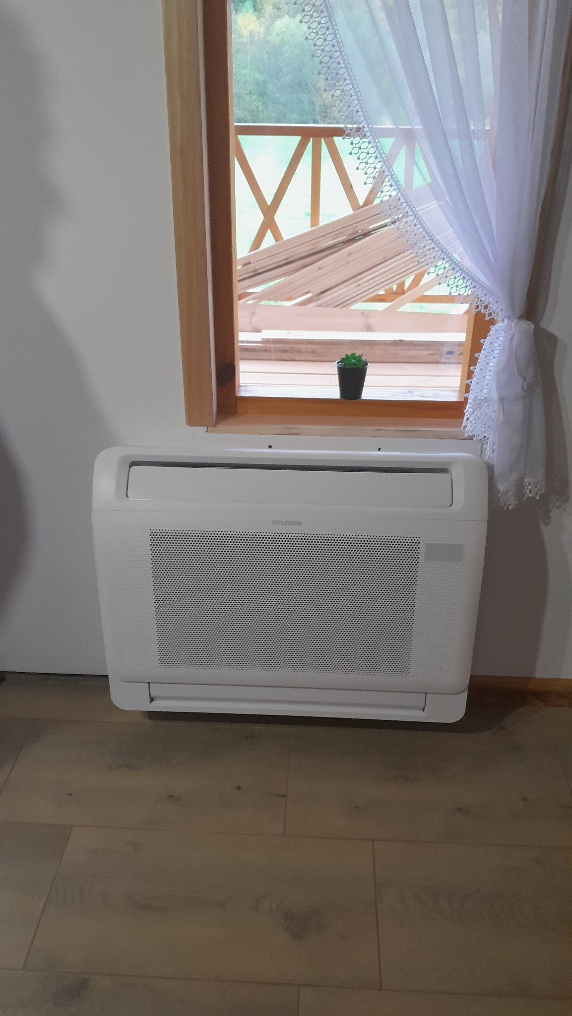 Biały klimatyzator marki Hyundai zamontowany w pomieszczeniu z drewnianą podłogą, widok na okno z drewnianą ramą, przez które widać niezabezpieczone deski na zewnątrz, okno ozdobione białą...