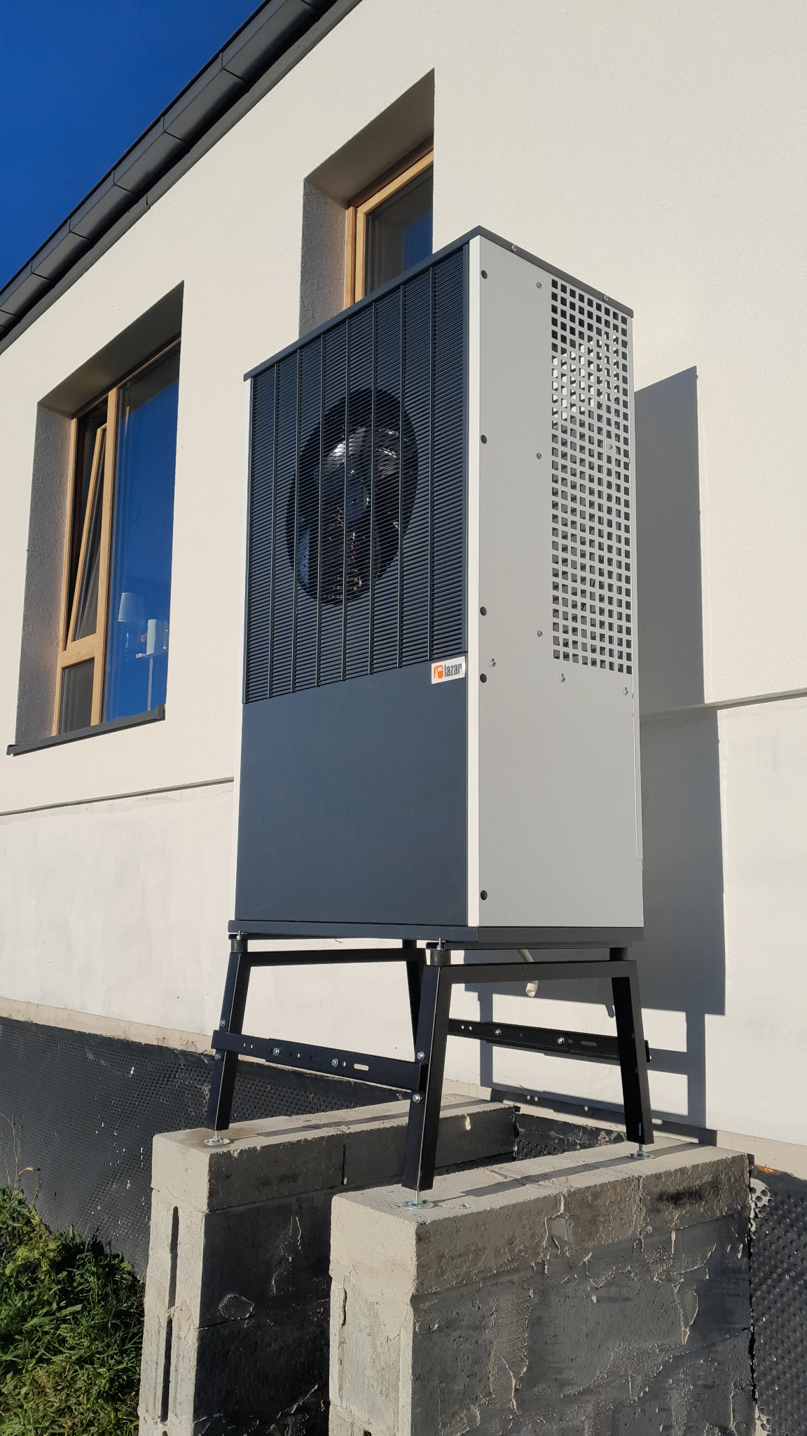 Zewnętrzna pompa ciepła powietrze-woda, model stojący na betonowych fundamentach przy białej ścianie budynku z widocznym oknem i niebieskim niebem w tle.