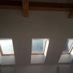 Trzy okna dachowe w nowo wykończonym poddaszu z drewnianymi belkami stropowymi, widoczne grzejniki pod oknami.