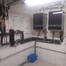 Dwa kotły grzewcze Buderus zamontowane na ścianie wyłożonej białymi płytkami, widoczne rury instalacji grzewczej i armatura, z niebieskim pojemnikiem pod spodem.