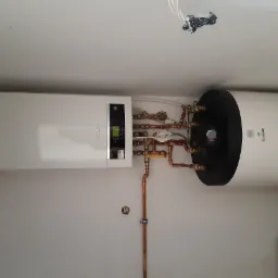 Nowoczesny, biały piec gazowy Buderus z podgrzewaczem wody Galmet, zamontowany na białej ścianie z widocznymi miedzianymi rurami i instalacją hydrauliczną, oraz niedokończonym gniazdkiem...