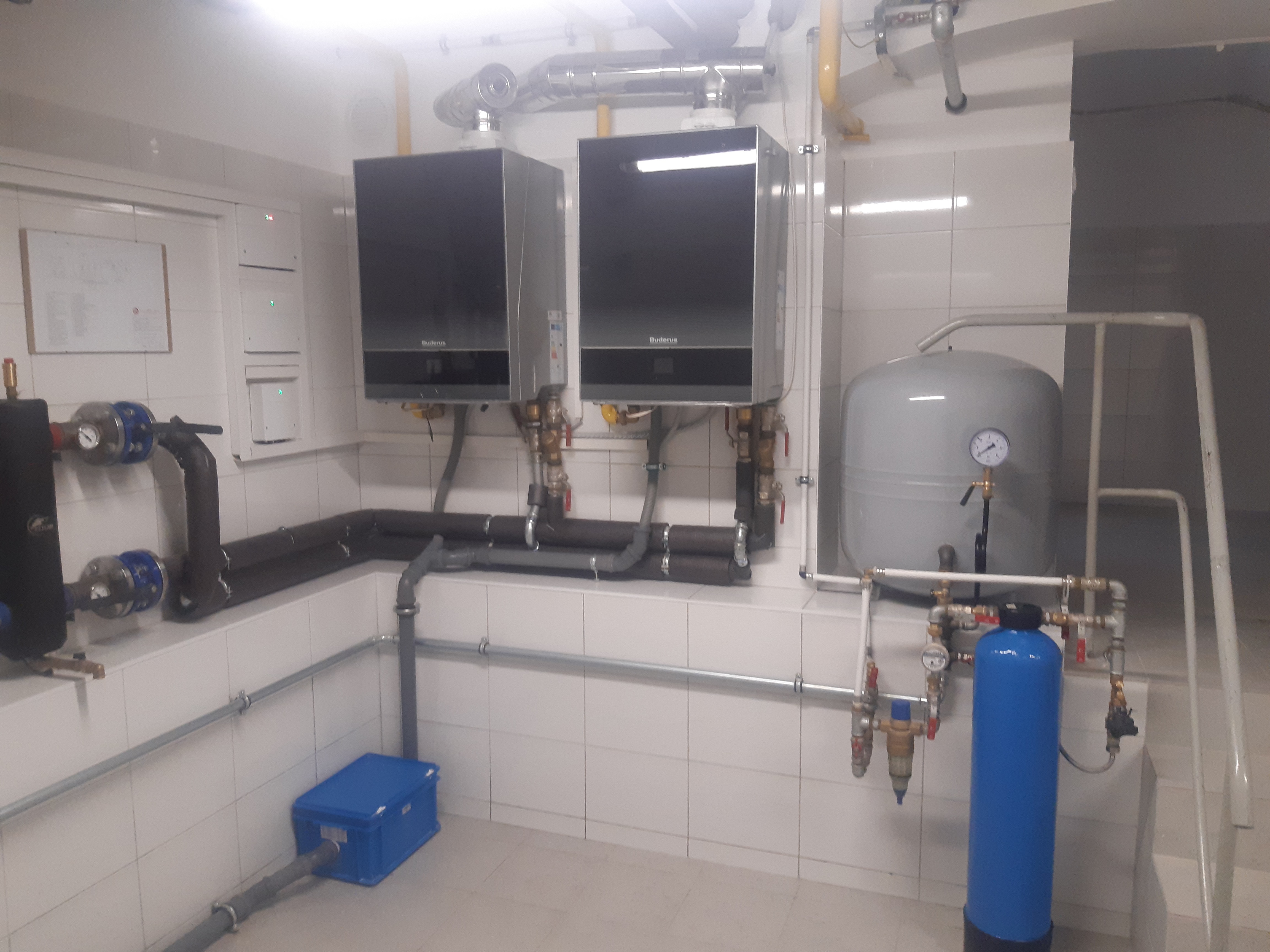 Dwa wiszące piece gazowe marki Buderus z podłączeniami, instalacja grzewcza z szarym zbiornikiem wyrównawczym i niebieskim filtrem wody, całość w pomieszczeniu z białymi płytkami.