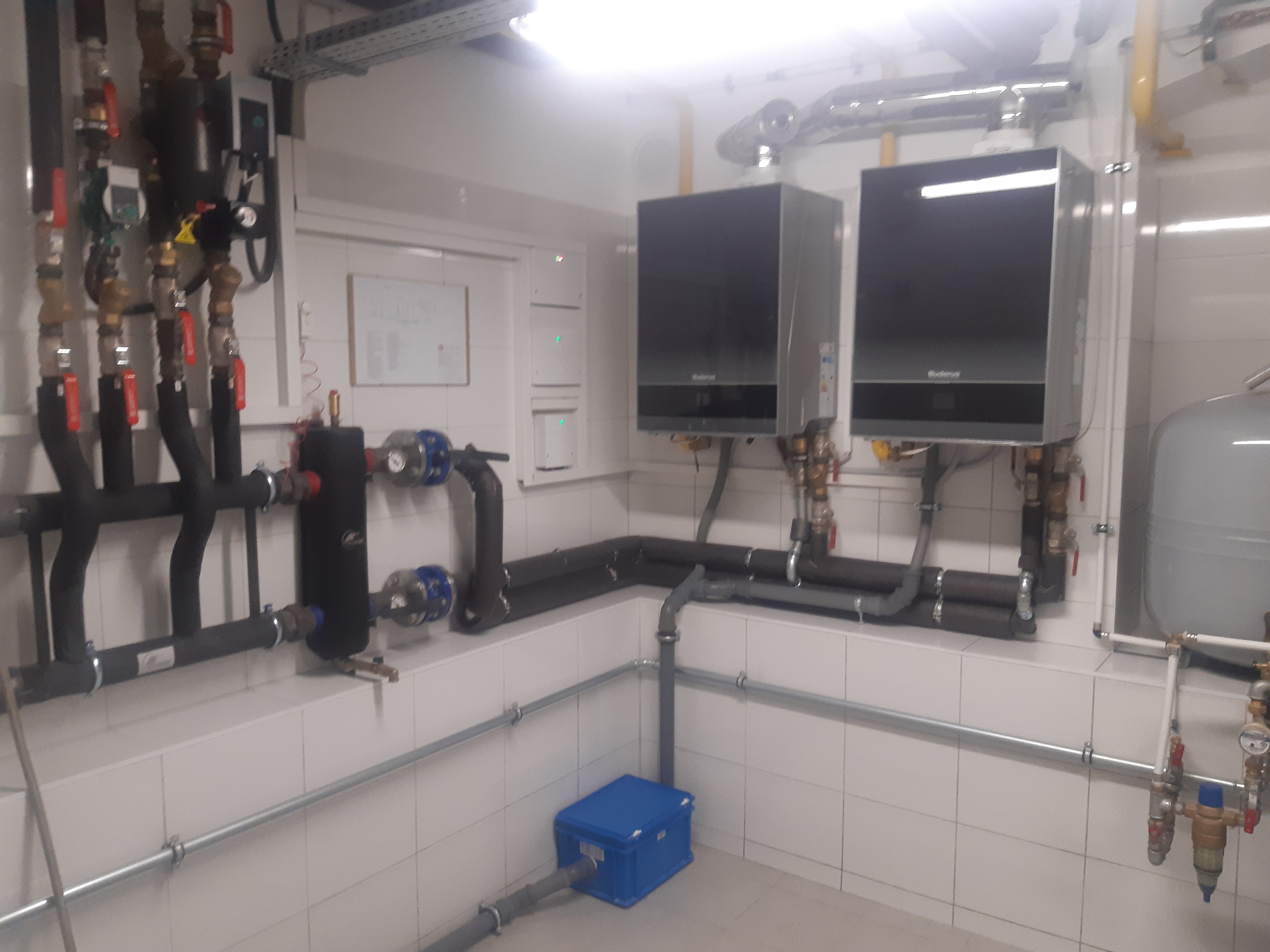 Dwa kotły grzewcze Buderus zamontowane na ścianie wyłożonej białymi płytkami, widoczne rury instalacji grzewczej i armatura, z niebieskim pojemnikiem pod spodem.