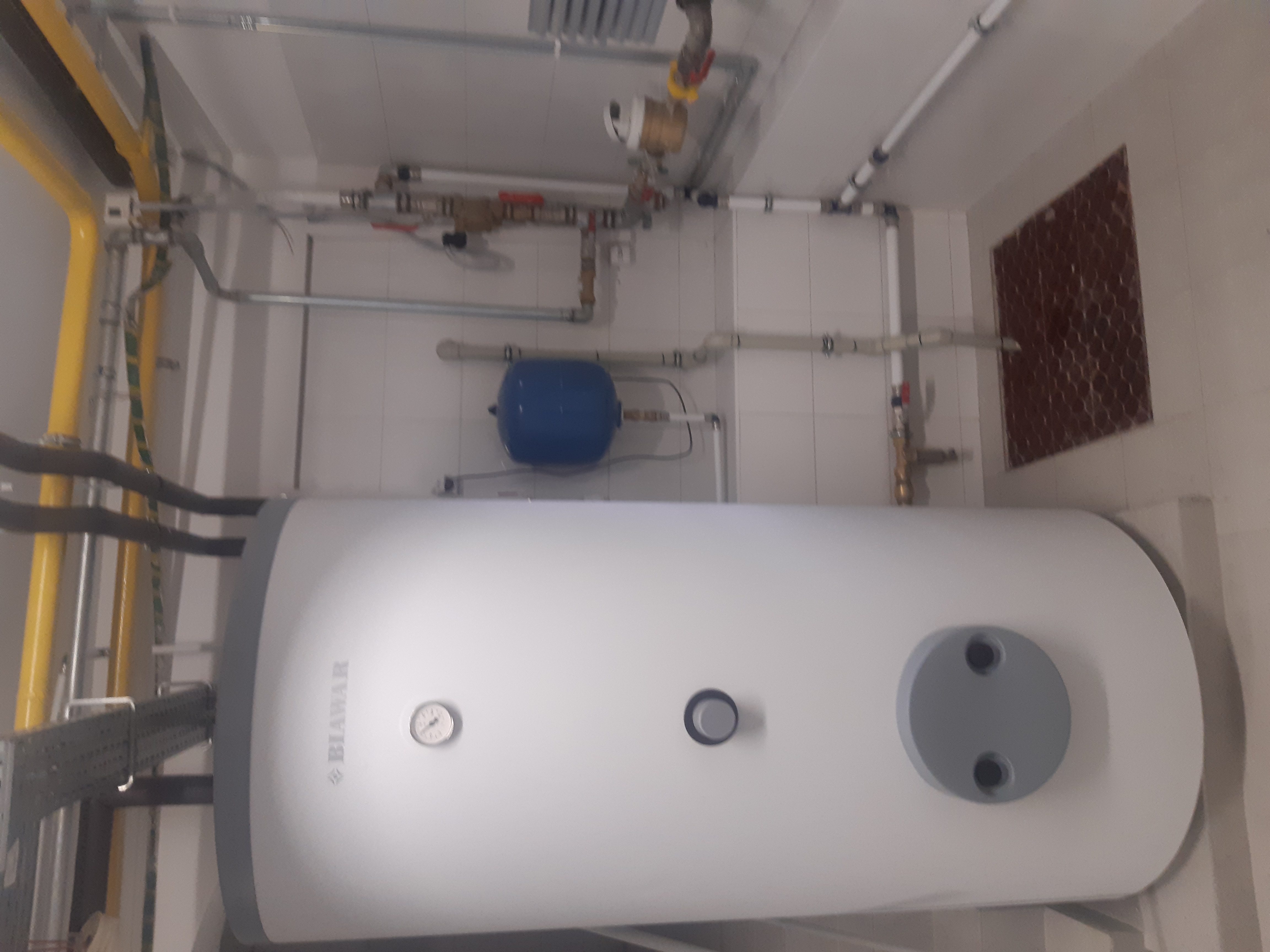 Biały bojler wiszący na ścianie z widocznymi rurami, zaworami, manometrem i niebieskim zbiornikiem wyrównawczym w pomieszczeniu z białymi płytkami.