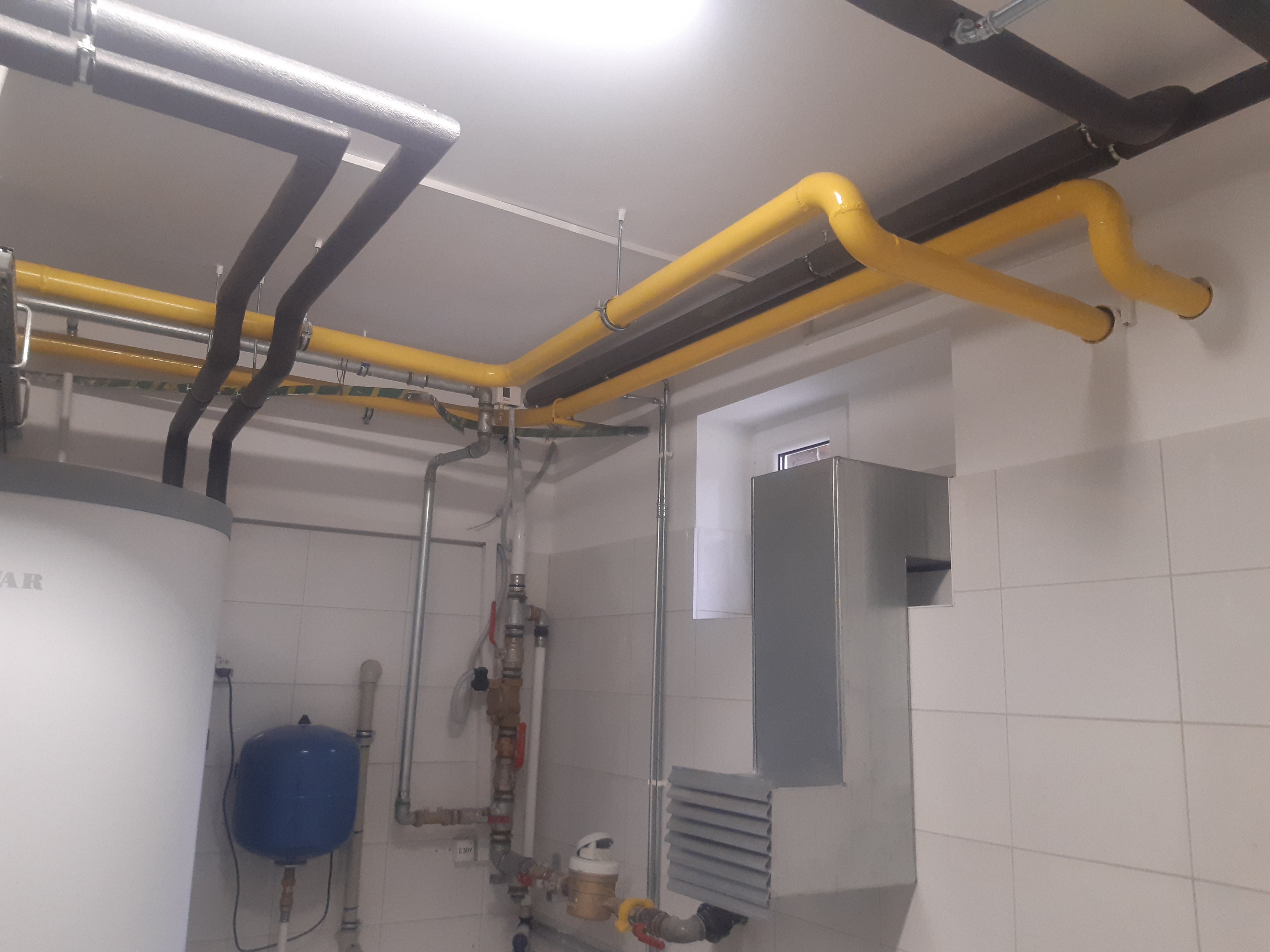 Wnętrze kotłowni z widocznymi rurami gazowymi w kolorze żółtym, rurami centralnego ogrzewania z izolacją termiczną, zbiornikiem ciśnieniowym w kolorze niebieskim i bojlerem, na ścianach białe płytki.