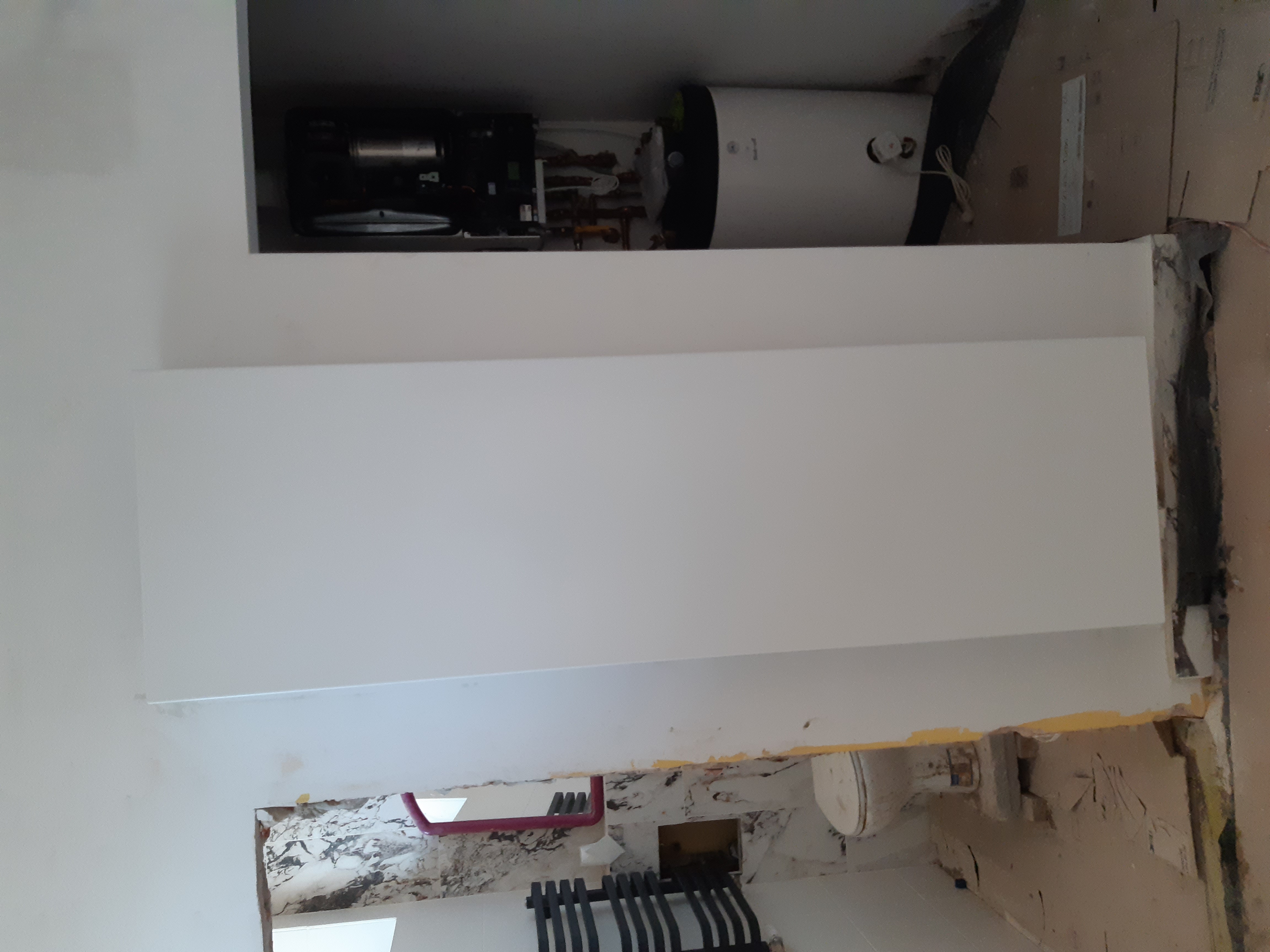 Biała zabudowa skrywająca piec gazowy dwufunkcyjny i zasobnik ciepłej wody, widoczne rury instalacji grzewczej i fragment grzejnika żeberkowego na tle remontowanego pomieszczenia.