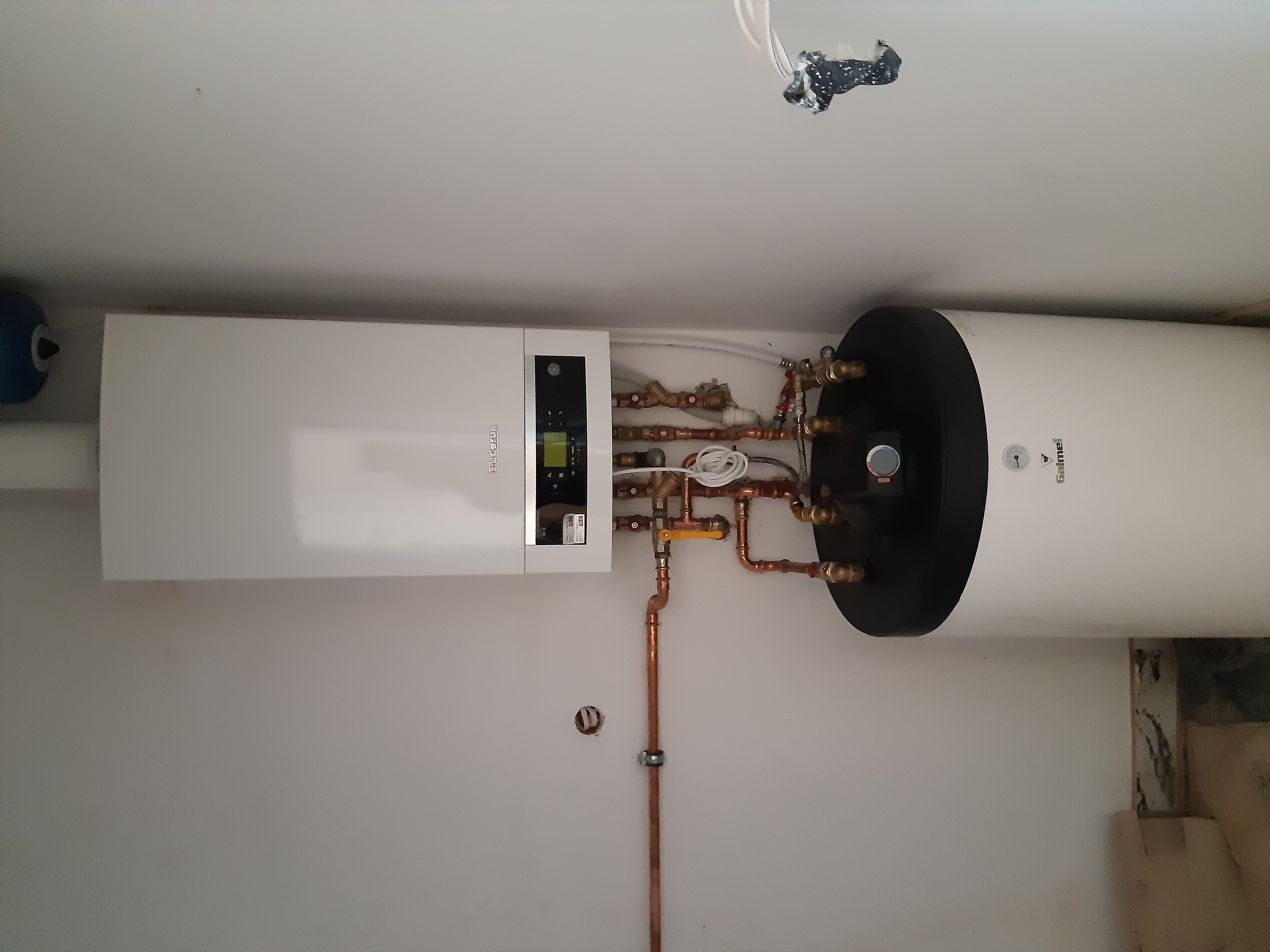Nowoczesny, biały piec gazowy Buderus z podgrzewaczem wody Galmet, zamontowany na białej ścianie z widocznymi miedzianymi rurami i instalacją hydrauliczną, oraz niedokończonym gniazdkiem...