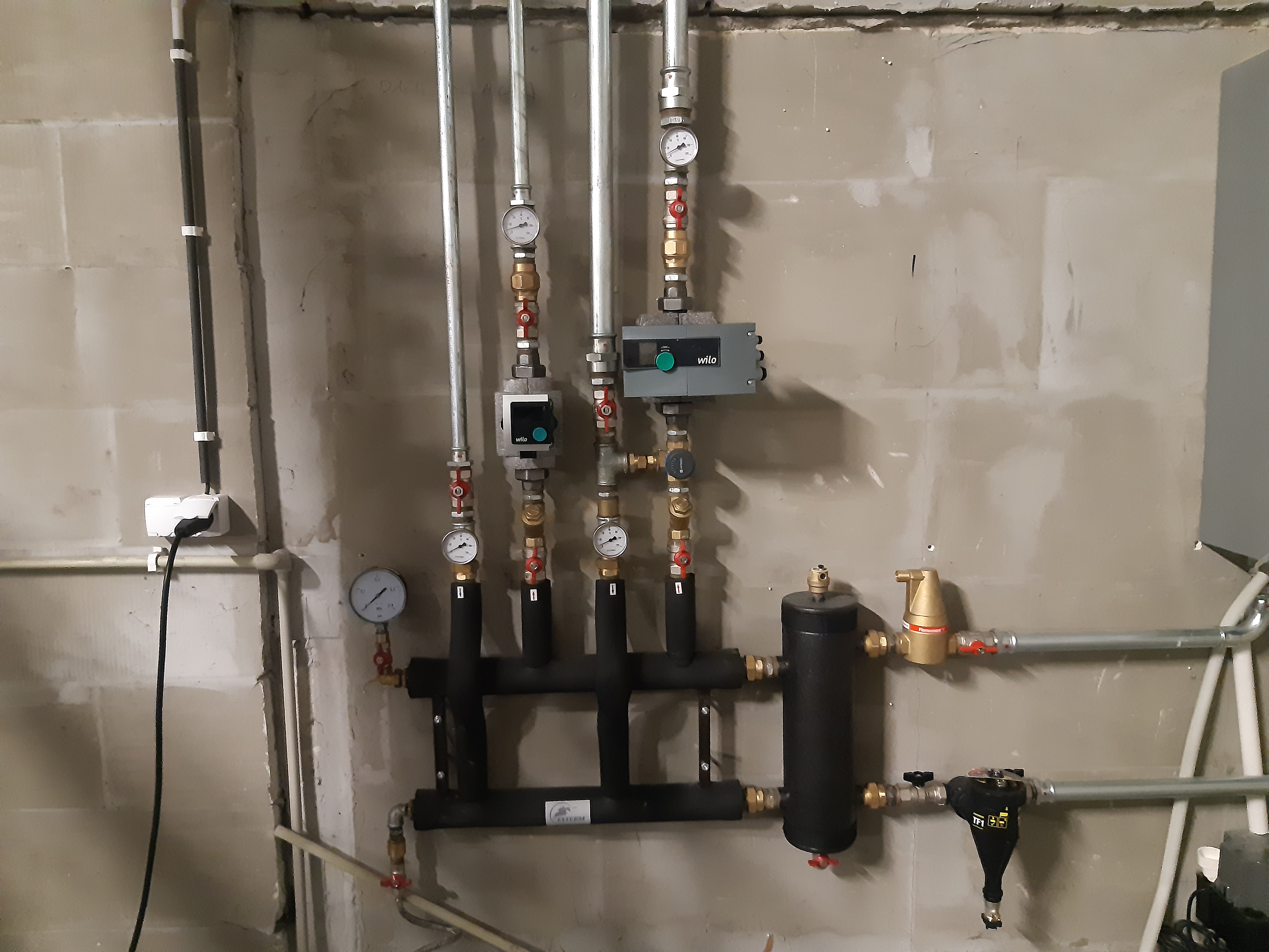 Rozdzielacz instalacji centralnego ogrzewania z manometrami i zaworami, zamontowany na surowej ścianie z widocznymi przewodami elektrycznymi.