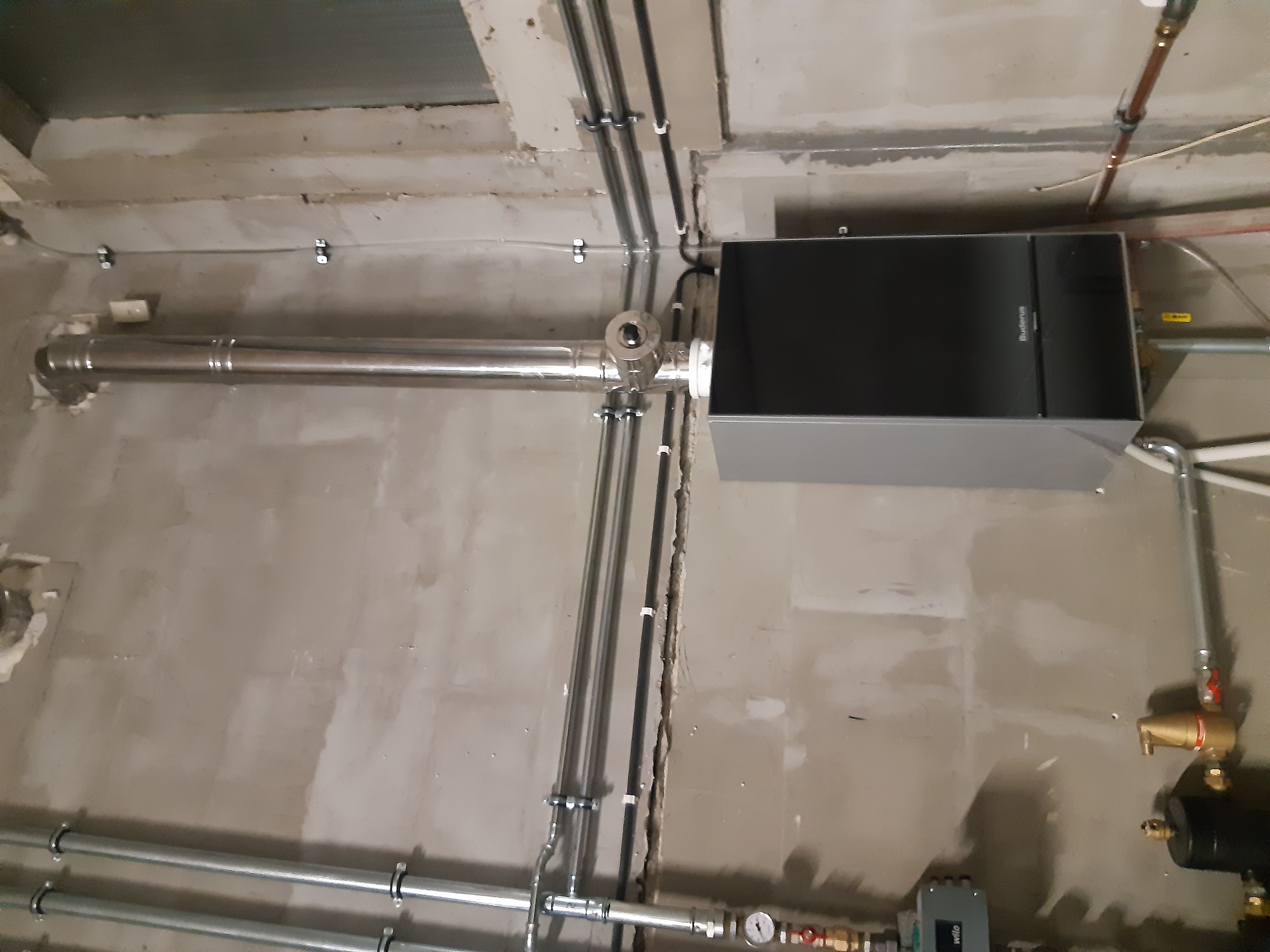 Zamontowany piec gazowy marki Buderus z orurowaniem na surowej ścianie z betonu komórkowego, widoczne czarne rury w peszlach i elementy instalacji grzewczej.