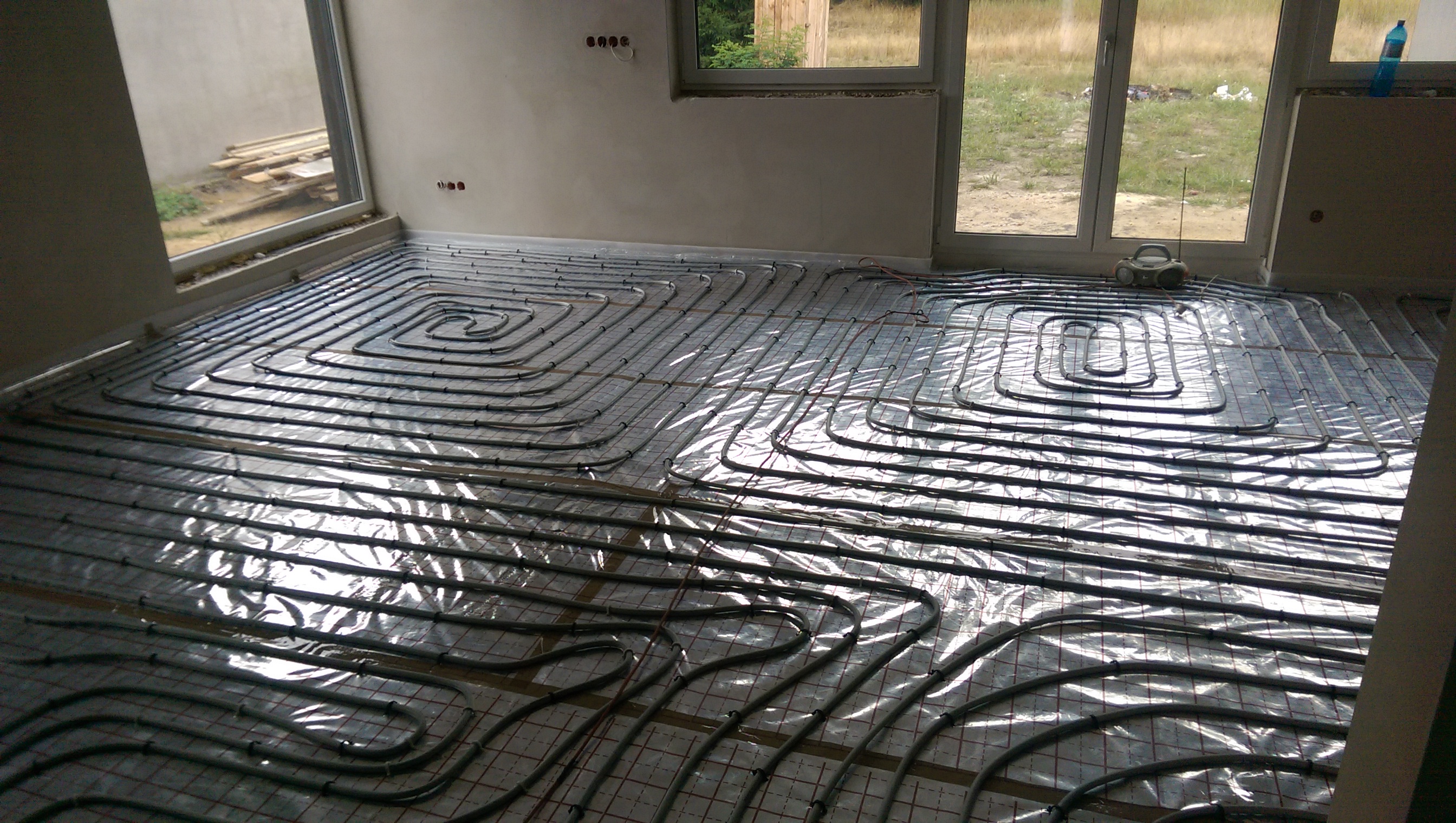 Rozłożone rury ogrzewania podłogowego na folii z siatką, widoczne okna i radio na parapecie w tle.