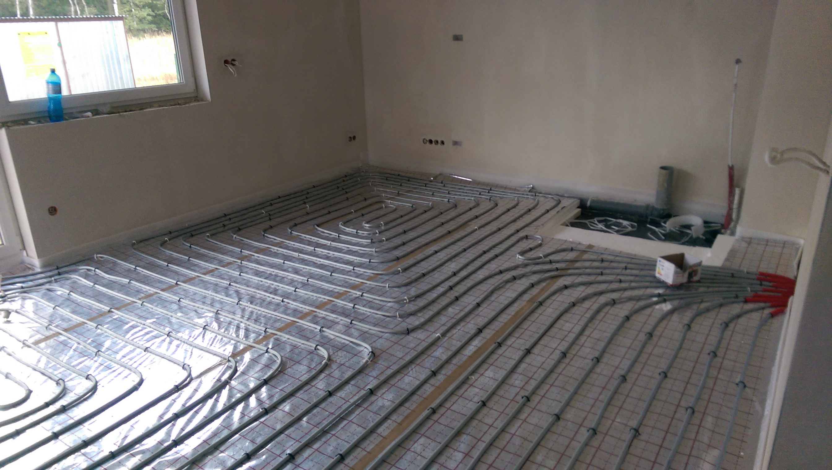 Szare rury ogrzewania podłogowego ułożone spiralnie na folii z siatką w nowym budynku, widoczne końcówki rur z czerwonymi zaślepkami i karton.