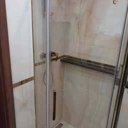 Krzysztof Kleparski - Kabina prysznicowa z złotymi profilami i armaturą, wykończona płytkami imitującymi marmur, z widoczną półką wewnątrz.