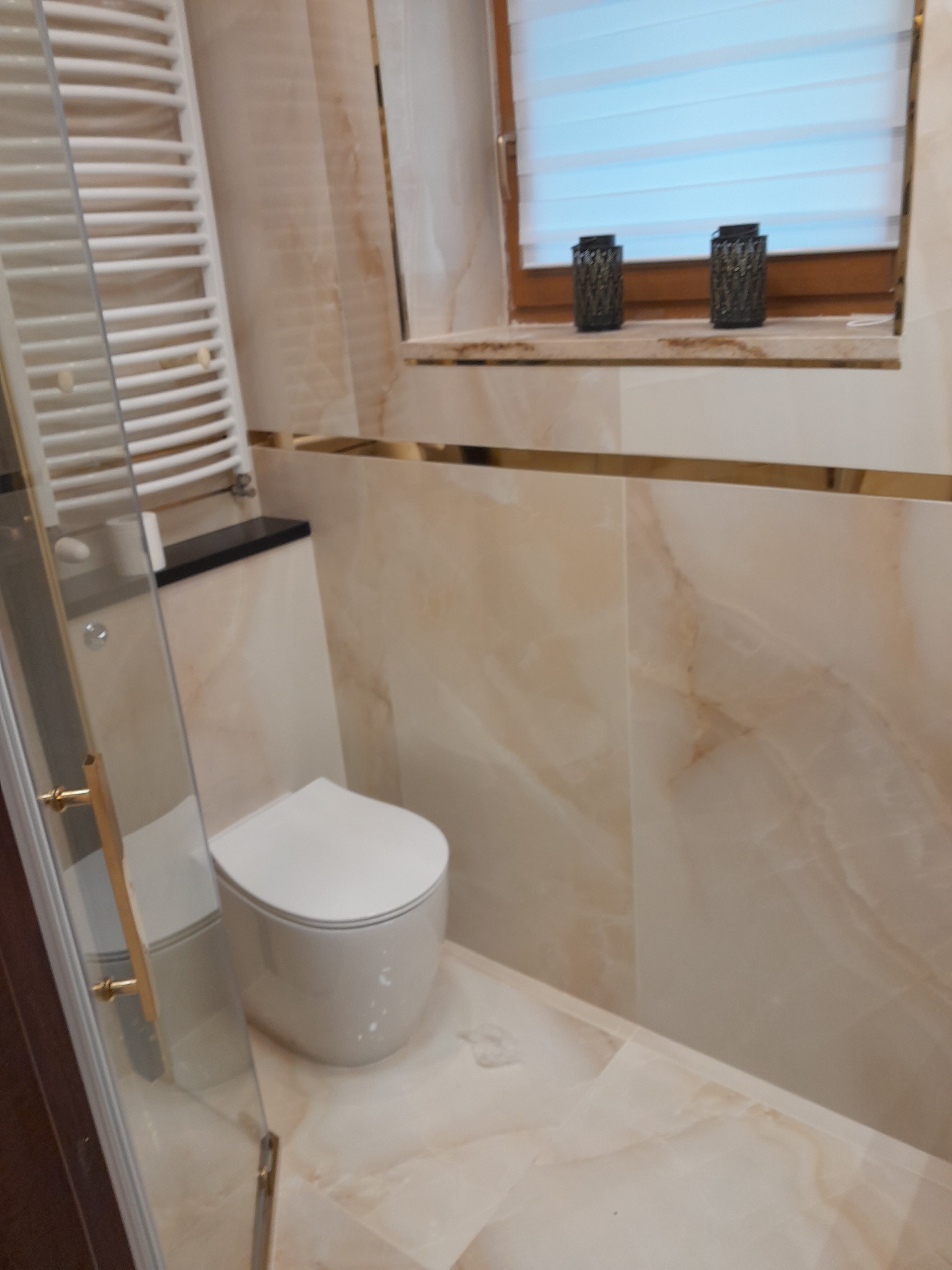 Nowoczesna łazienka z toaletą typu rimless, grzejnikiem drabinkowym i marmurkowymi płytkami w odcieniach beżu, doświetlona przez okno z żaluzjami.