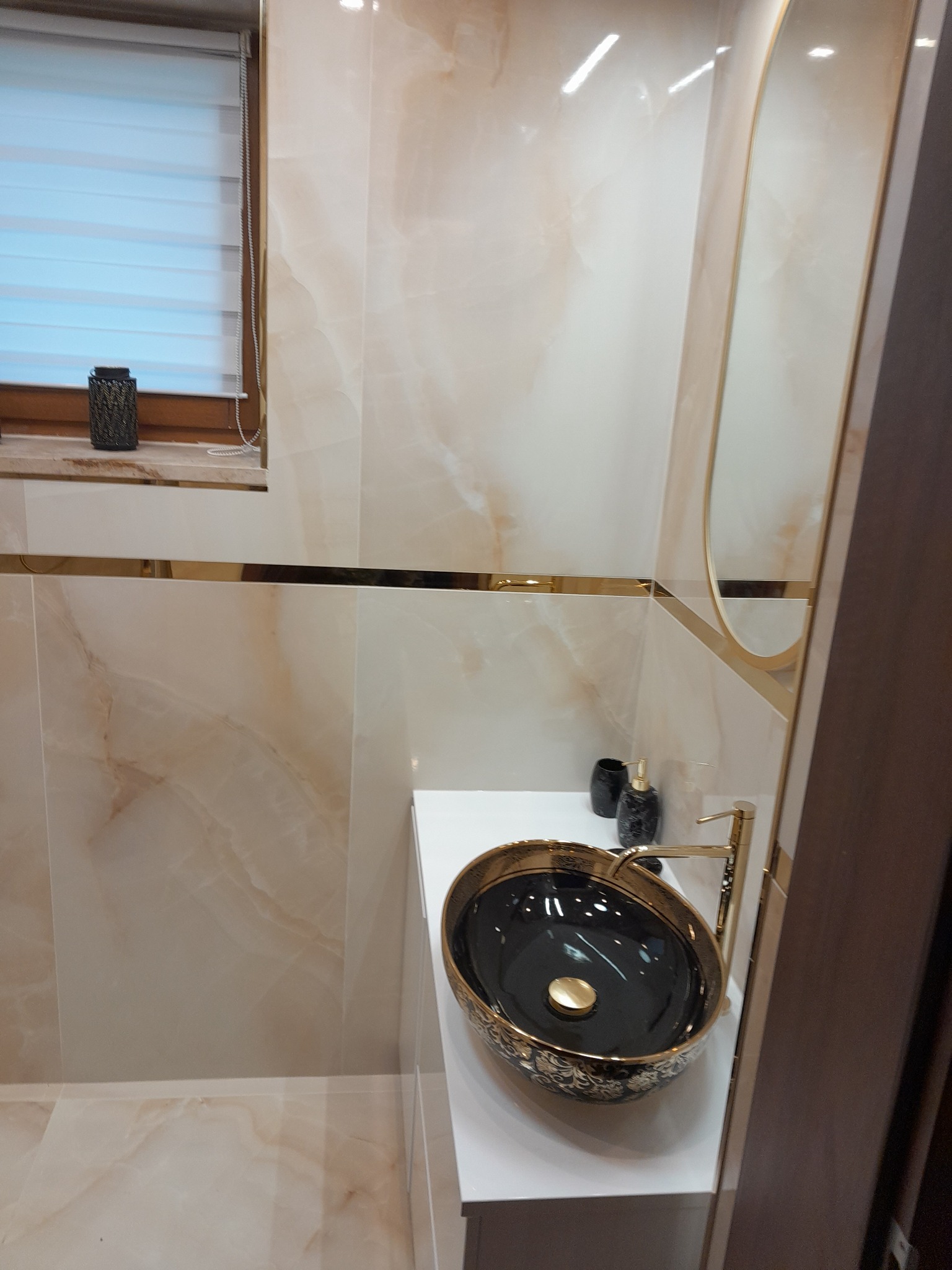 Elegancka łazienka z beżowymi marmurkowymi płytkami, złotymi akcentami i umywalką w stylu glamour z czarnym wnętrzem i złotymi zdobieniami.