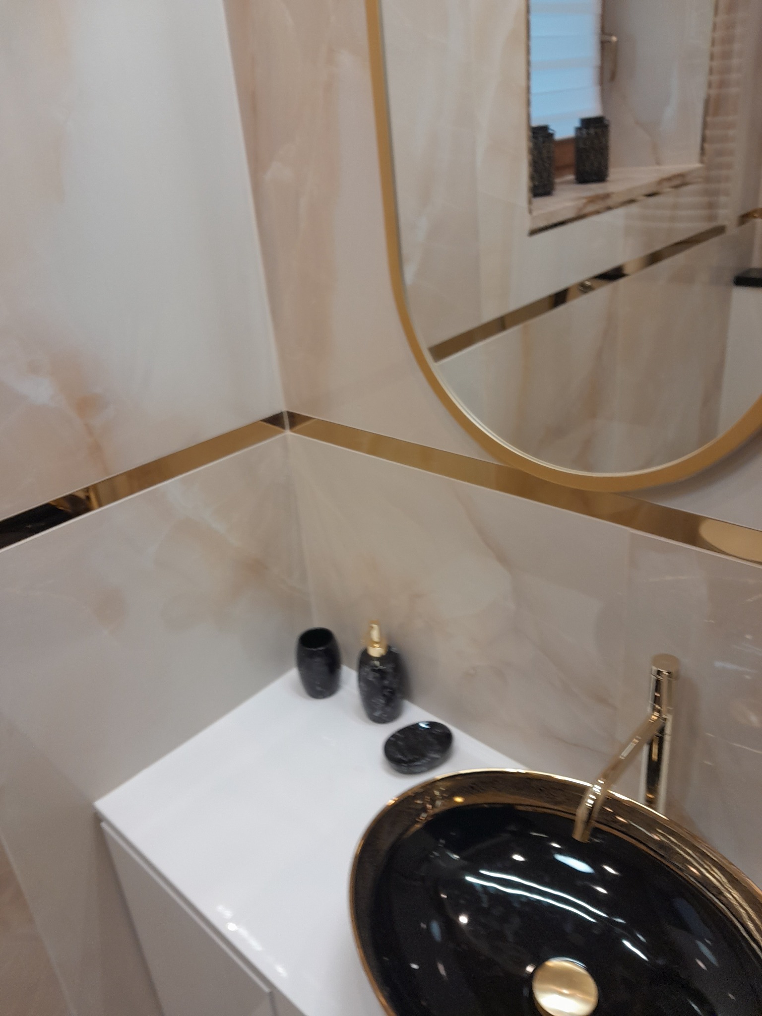Błyszcząca czarna umywalka z złotą armaturą na białej szafce, uzupełniona o marmurowe płytki i złote akcenty wykończeniowe w nowoczesnej łazience.