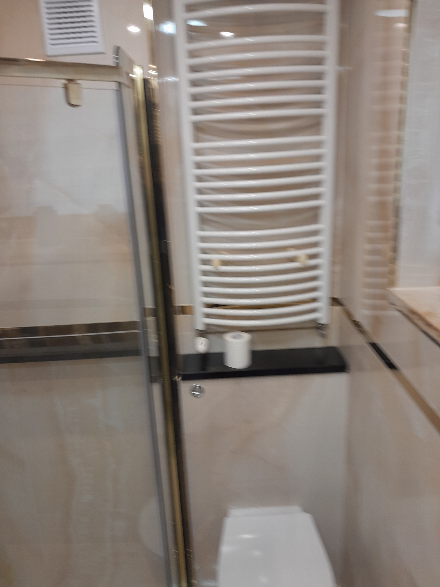 Nowoczesna łazienka z kabiną prysznicową ze złotymi ramami, grzejnikiem drabinkowym i toaletą w jasnych, beżowych odcieniach.