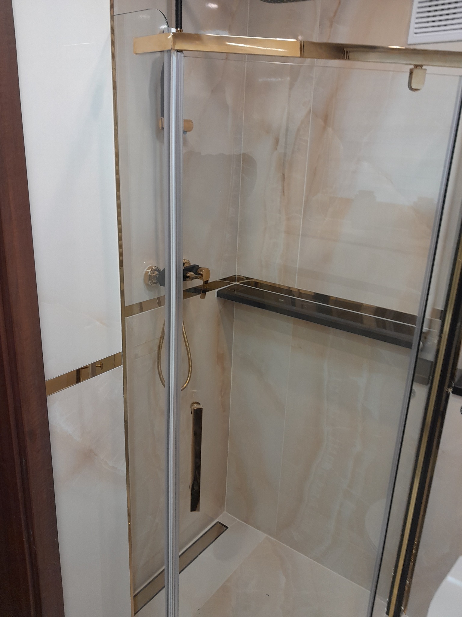 Kabina prysznicowa z złotymi profilami i armaturą, wykończona płytkami imitującymi marmur, z widoczną półką wewnątrz.