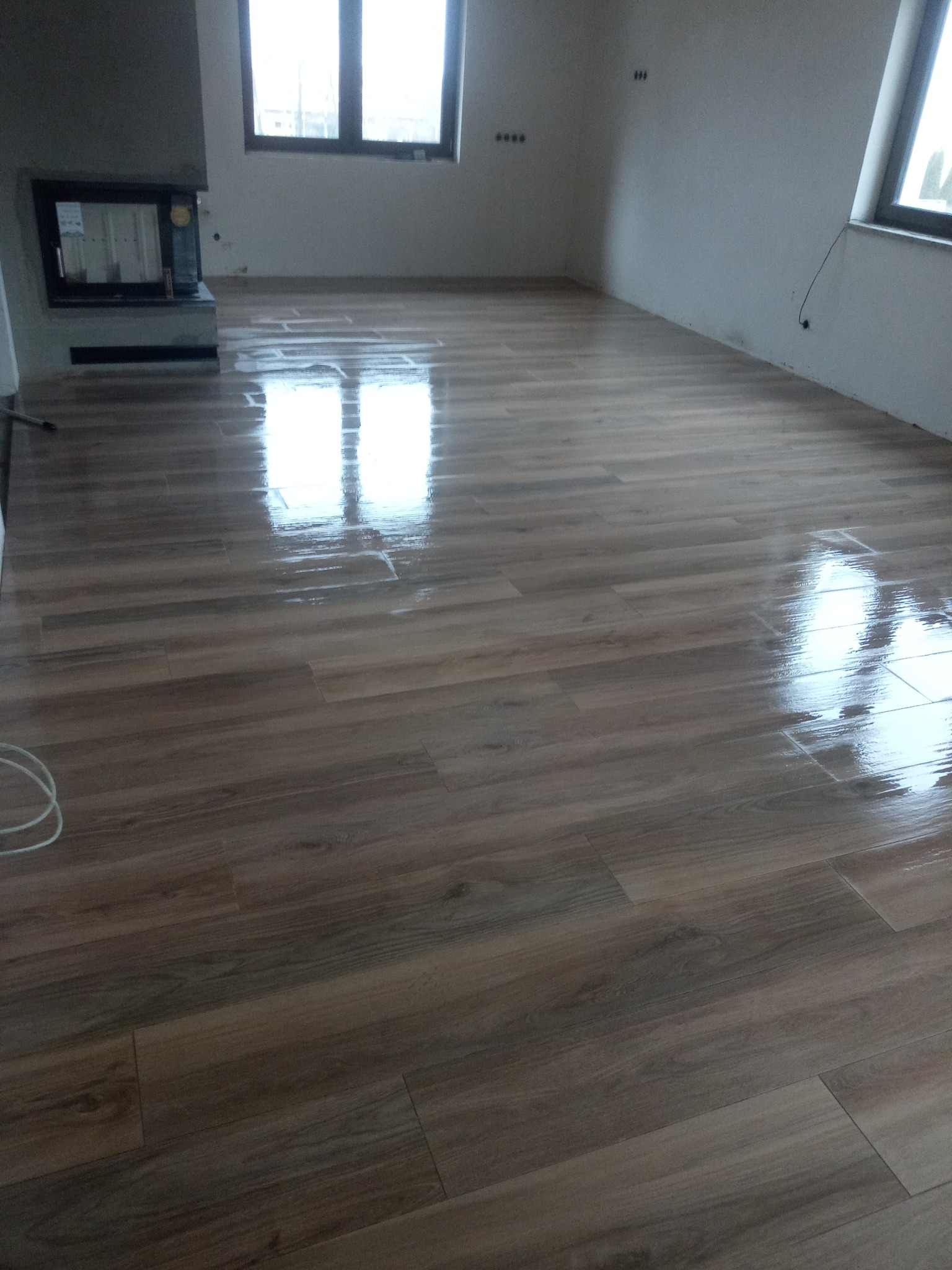 Ułożona podłoga z paneli imitujących drewno, z widocznymi refleksami światła, w surowym wnętrzu z oknami i kominkiem.