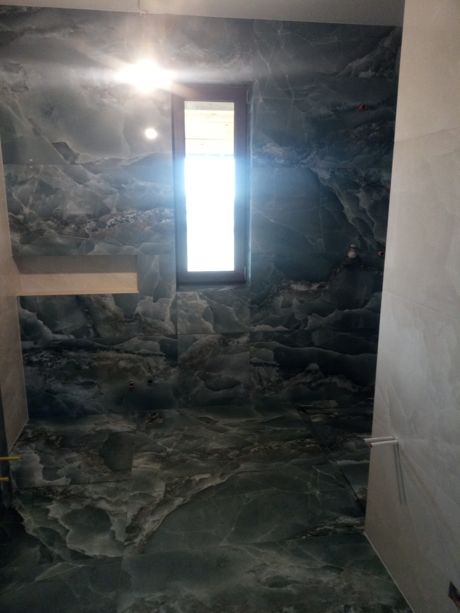 Wykończona łazienka z ciemnymi płytkami imitującymi marmur na ścianach i podłodze, widoczne okno z jasnym światłem dziennym oraz fragment białej ściany z zamontowanym wieszakiem na ręczniki.