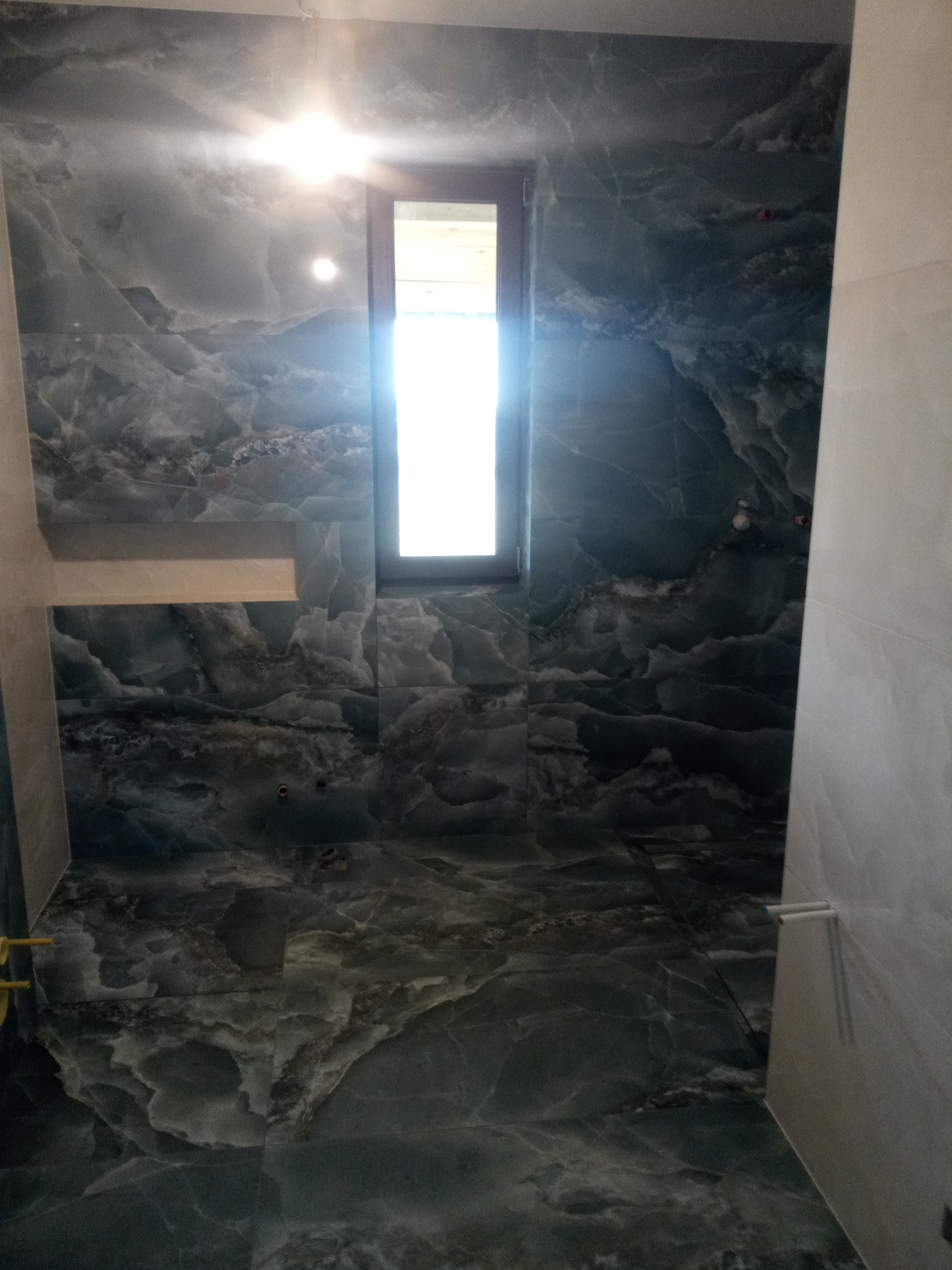 Wykończona łazienka z ciemnozielonymi płytkami imitującymi marmur na ścianach i podłodze, małe okno w ciemnej ramie, widoczny odpływ liniowy.