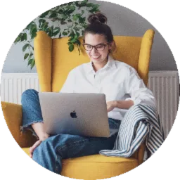 Uśmiechnięta kobieta w okularach siedzi w żółtym fotelu z laptopem na kolanach, w tle widoczna roślina doniczkowa.