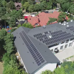 Widok z lotu ptaka na budynek z szarym dachem pokrytym panelami słonecznymi, otoczony zielenią drzew i zabudową mieszkalną.