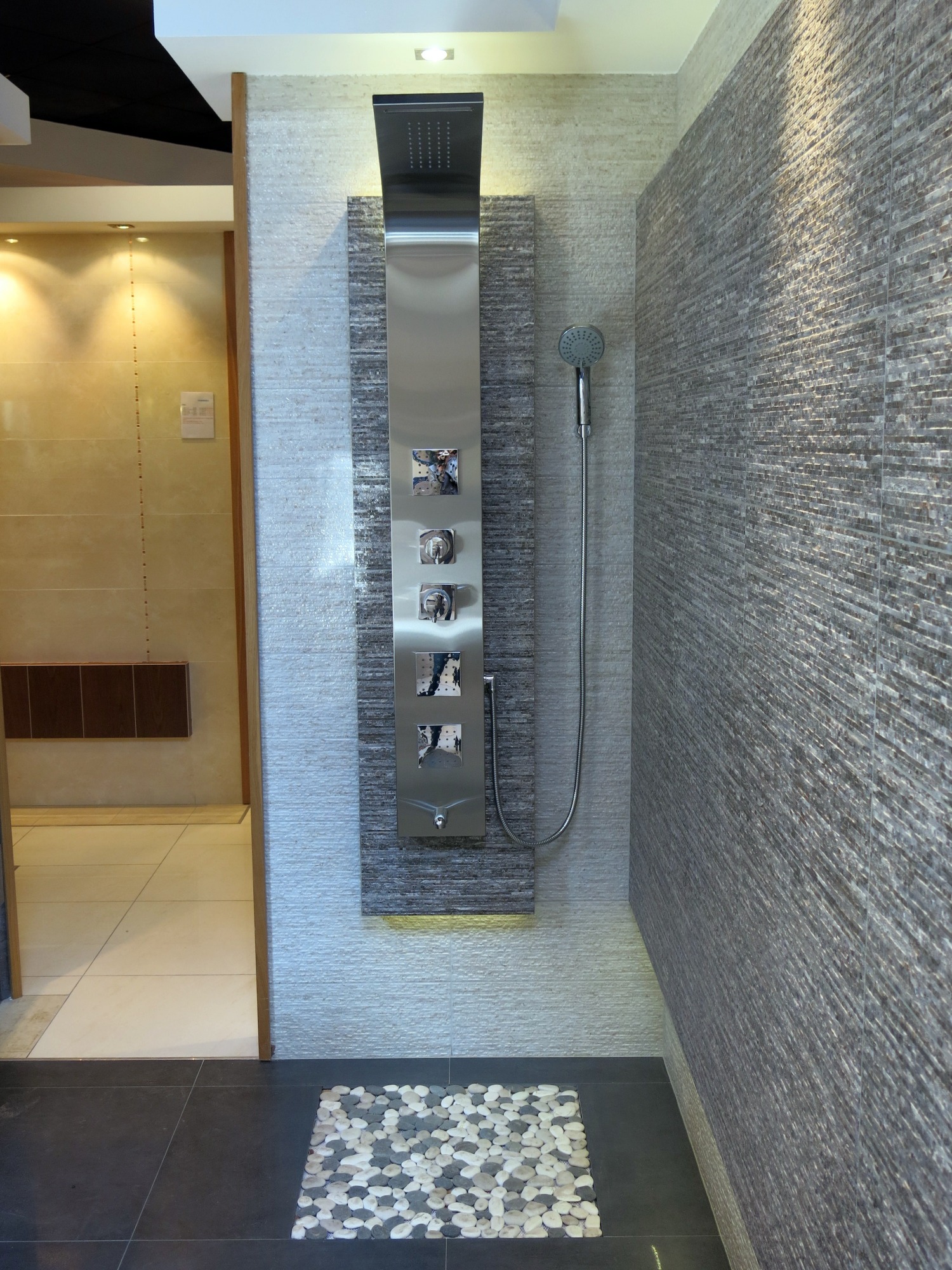Nowoczesna kabina prysznicowa z panelem natryskowym ze stali nierdzewnej i deszczownicą, wykończona płytkami imitującymi kamień, z dekoracyjnym dywanikiem z otoczaków na podłodze.