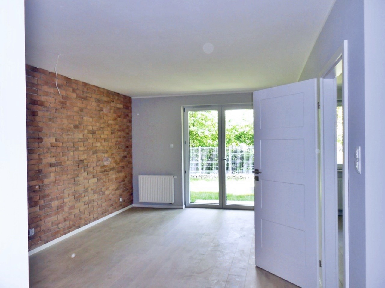 Jasne, puste pomieszczenie z jedną ścianą z cegły, białą podłogą, grzejnikiem i otwartymi białymi drzwiami balkonowymi prowadzącymi na zewnątrz.