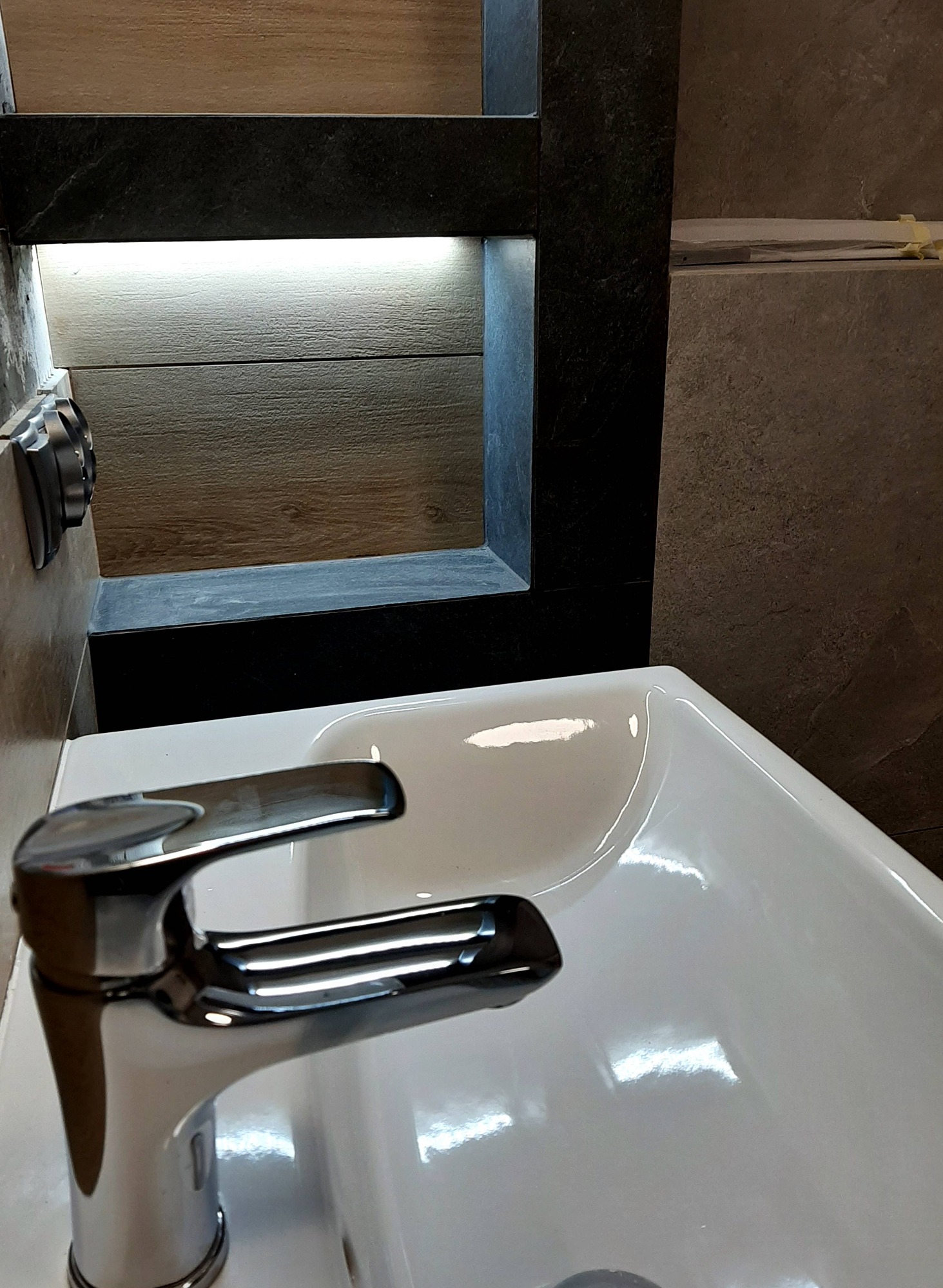 Biała umywalka z chromowaną baterią na tle wykończonej ściany z podświetlaną wnęką w łazience.