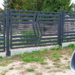 Nowoczesne, szare ogrodzenie metalowe z poziomymi panelami i geometrycznym wzorem w Skarszewach, osadzone na betonowej podmurówce.
