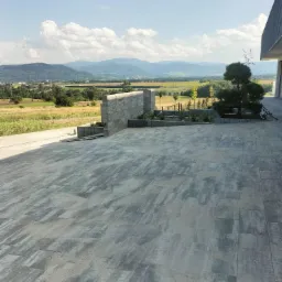 Szara kostka brukowa ułożona na dużym placu przed nowoczesnym domem z widokiem na góry i pola w tle, elementem dekoracyjnym jest mur z kostki i zasadzone rośliny.