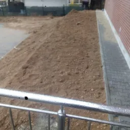 Nierówny teren z nasypem ziemi przy ścianie budynku z kostką brukową, widziany zza metalowego ogrodzenia.