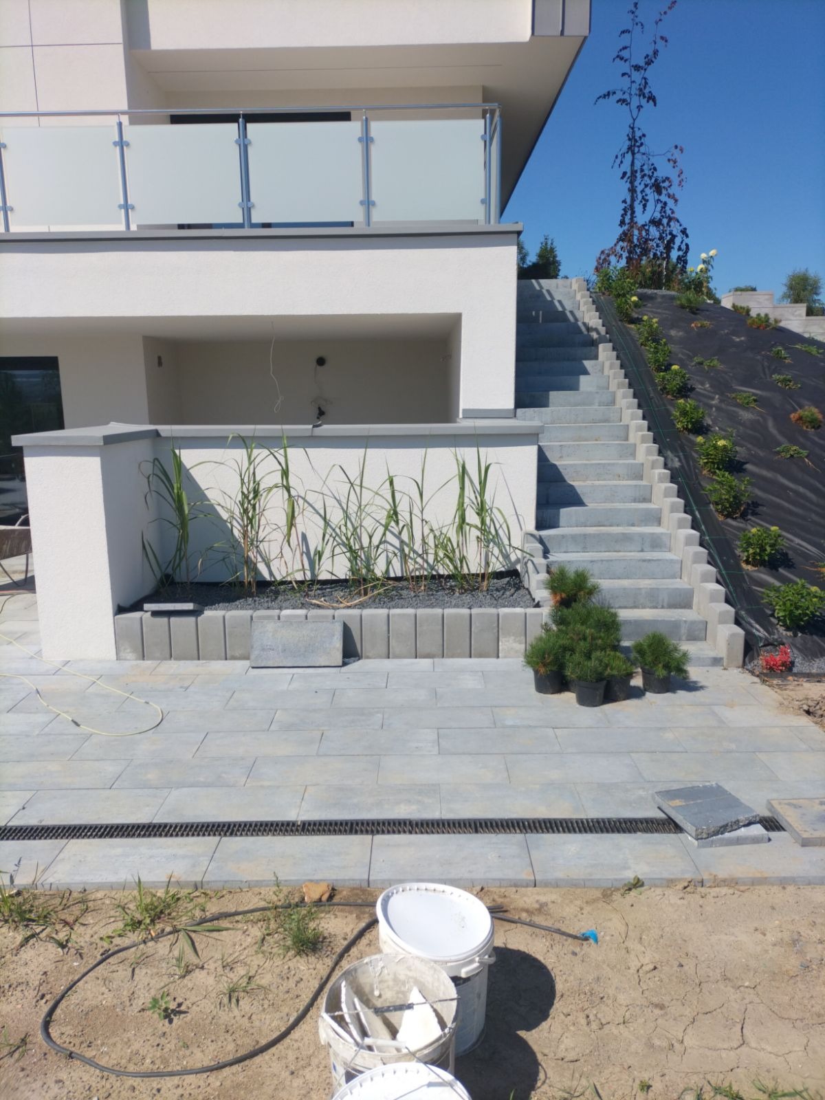 Nowoczesny dom z tarasem i schodami wyłożonymi szarą kostką brukową, widoczne doniczki z iglakami, w tle skarpa zabezpieczona czarną agrowłókniną, na pierwszym planie wiaderka po farbie.