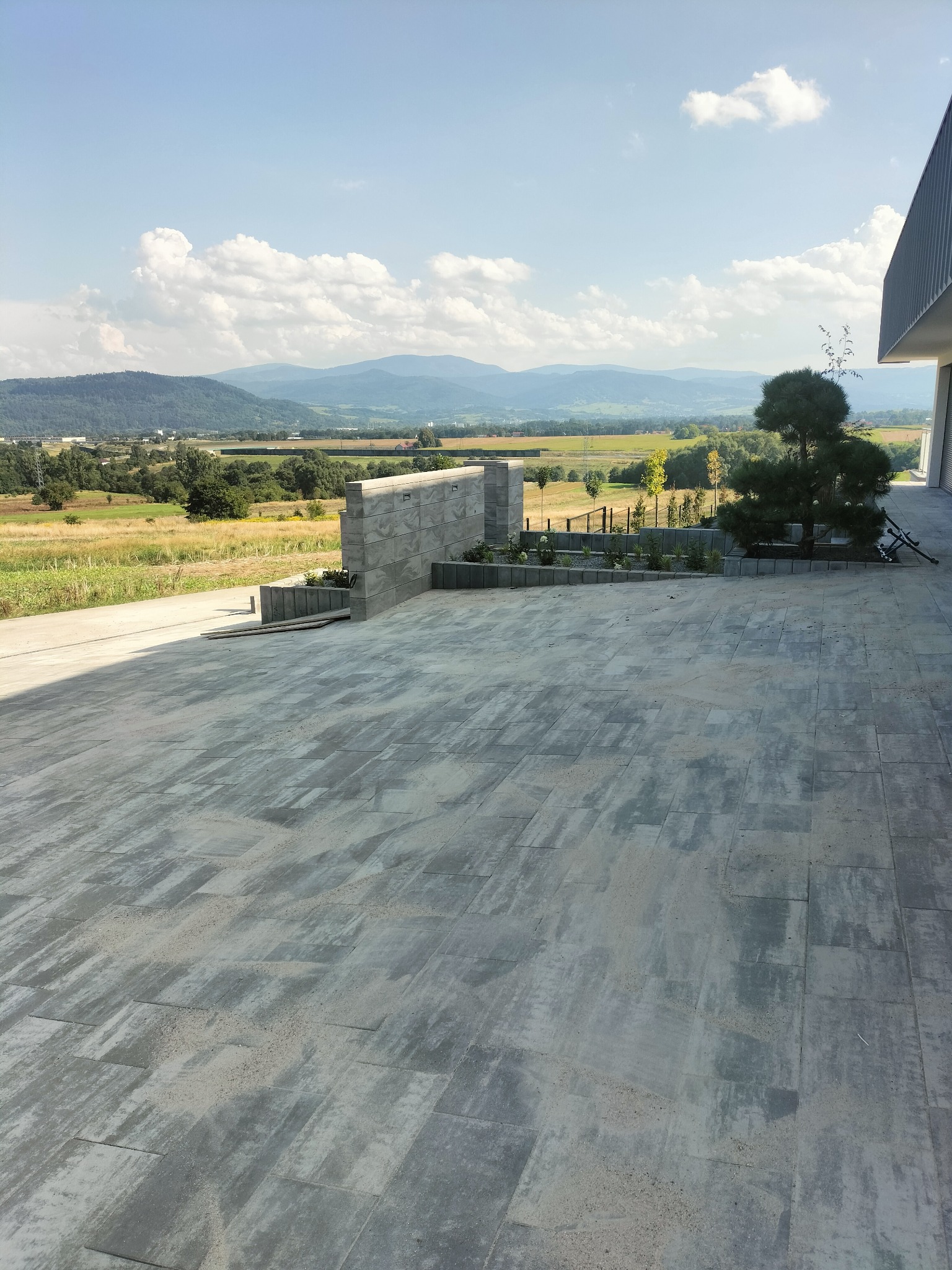 Szara kostka brukowa ułożona na dużym placu przed nowoczesnym domem z widokiem na góry i pola w tle, elementem dekoracyjnym jest mur z kostki i zasadzone rośliny.