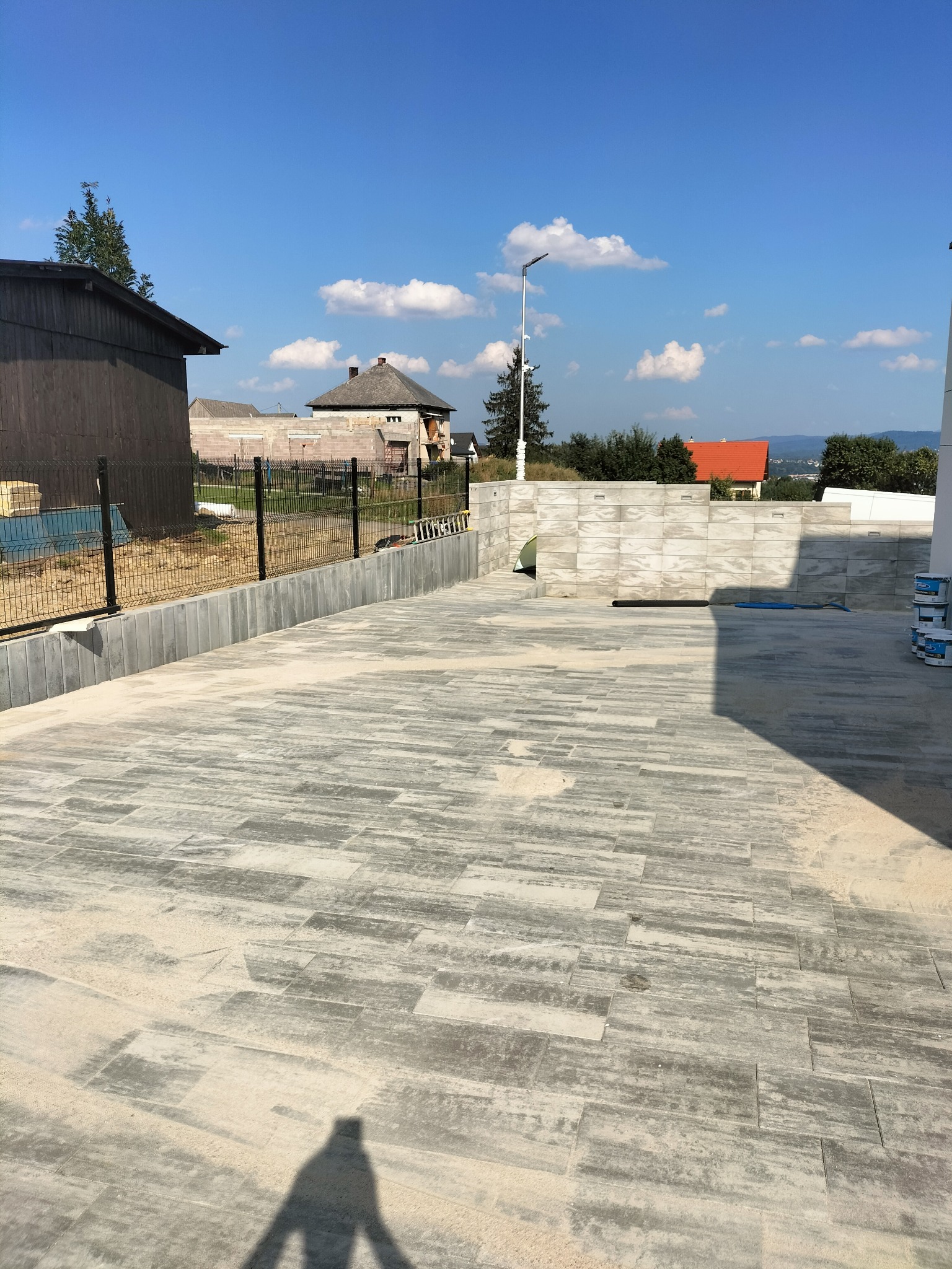 Ułożona, szara kostka brukowa na posesji z widocznym ogrodzeniem panelowym, budynkami w tle i shadow użytkownika.